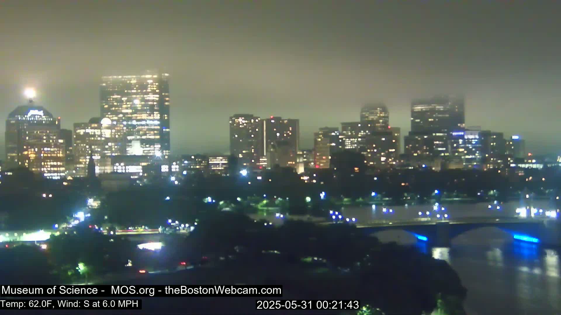 A nighttime cityscape, shrouded in fog, shows a city skyline with illuminated buildings and a bridge over a body of water.