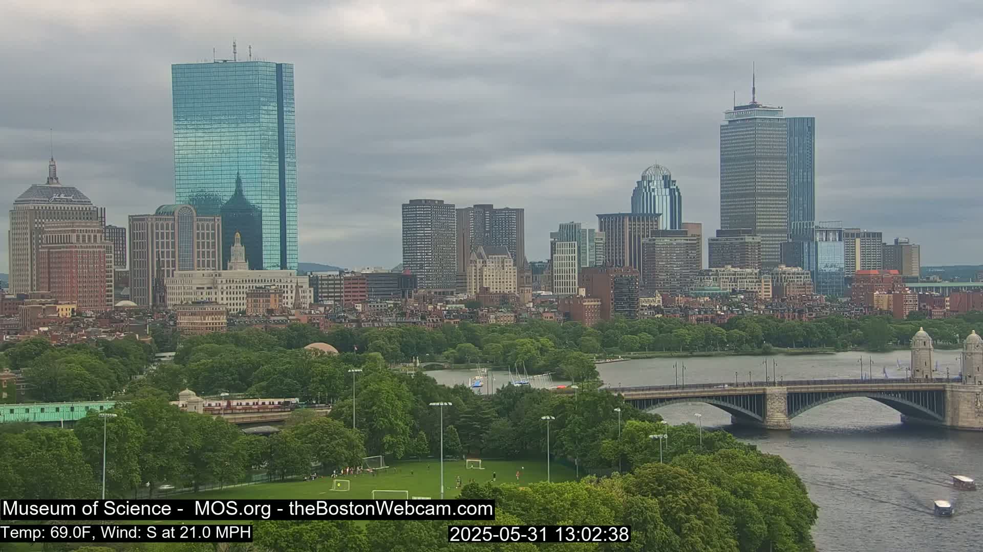 A city skyline under a cloudy sky is seen from across a river and a bridge, with green parkland in the foreground.