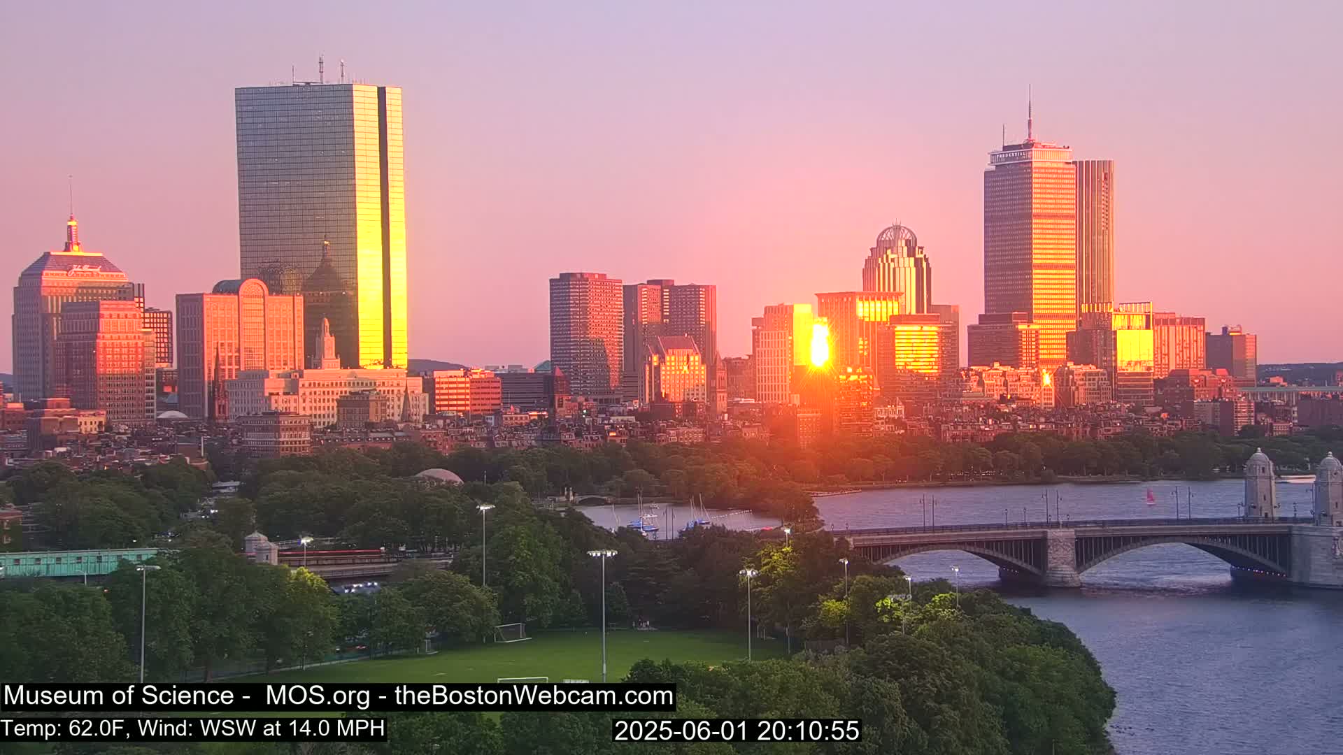 A city skyline at sunset, with the sun setting behind several tall buildings, viewed from across a river with a bridge and green space in the foreground.