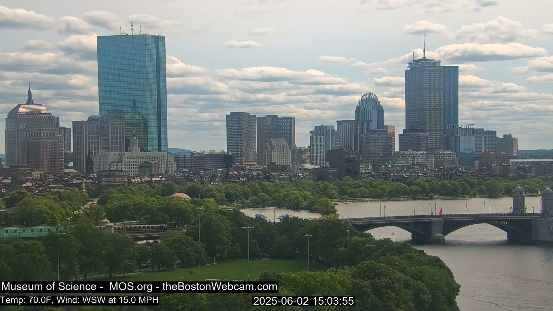 A city skyline with numerous skyscrapers and buildings is visible across a river with a bridge, under a partly cloudy sky.