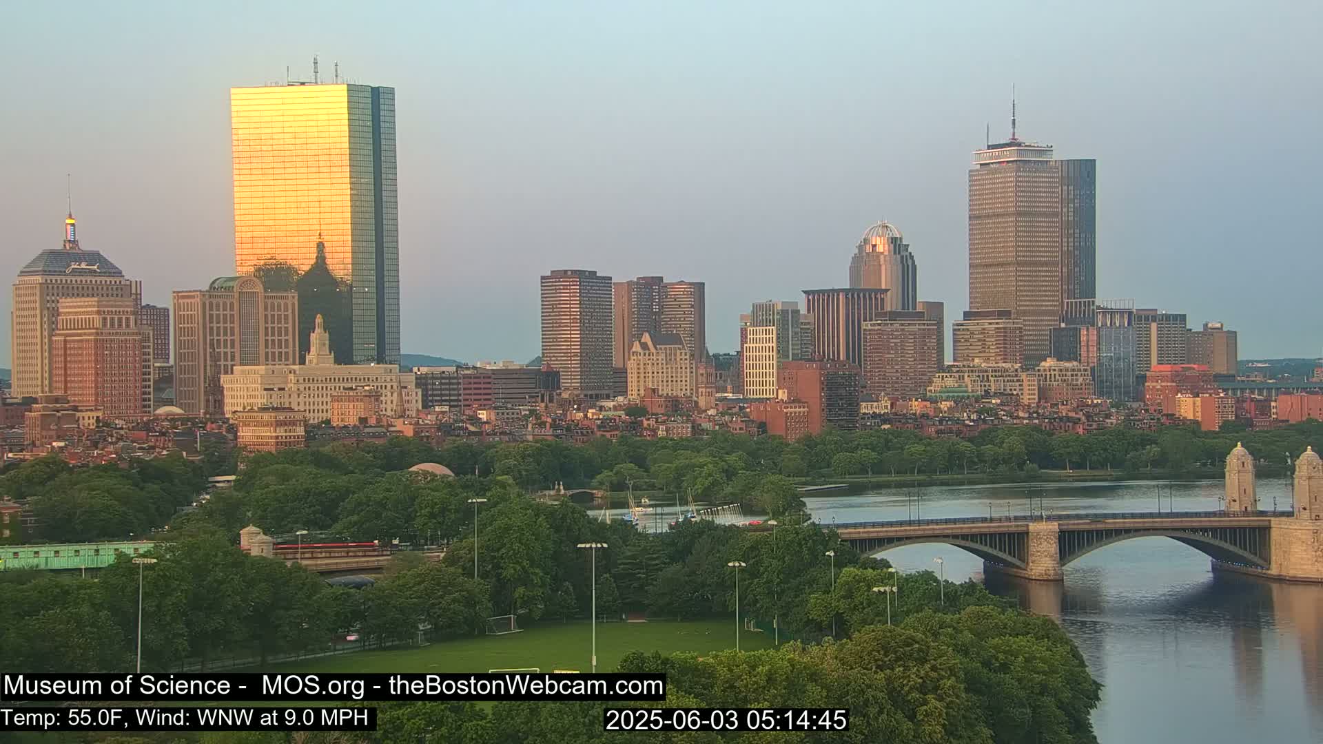 A cityscape at sunrise, showing a river with a bridge and many buildings, under a mostly clear sky.