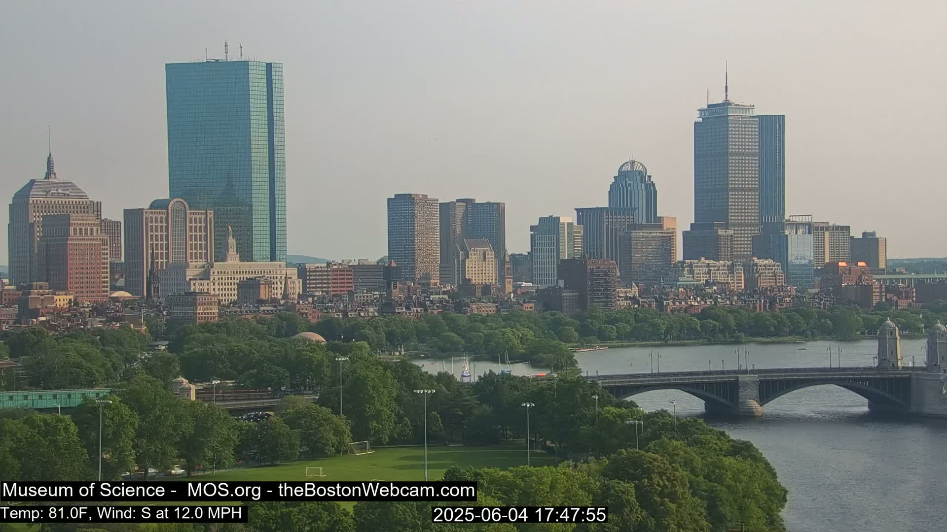 A city skyline with numerous skyscrapers is visible across a river, with a bridge in the foreground and a calm, hazy sky above.