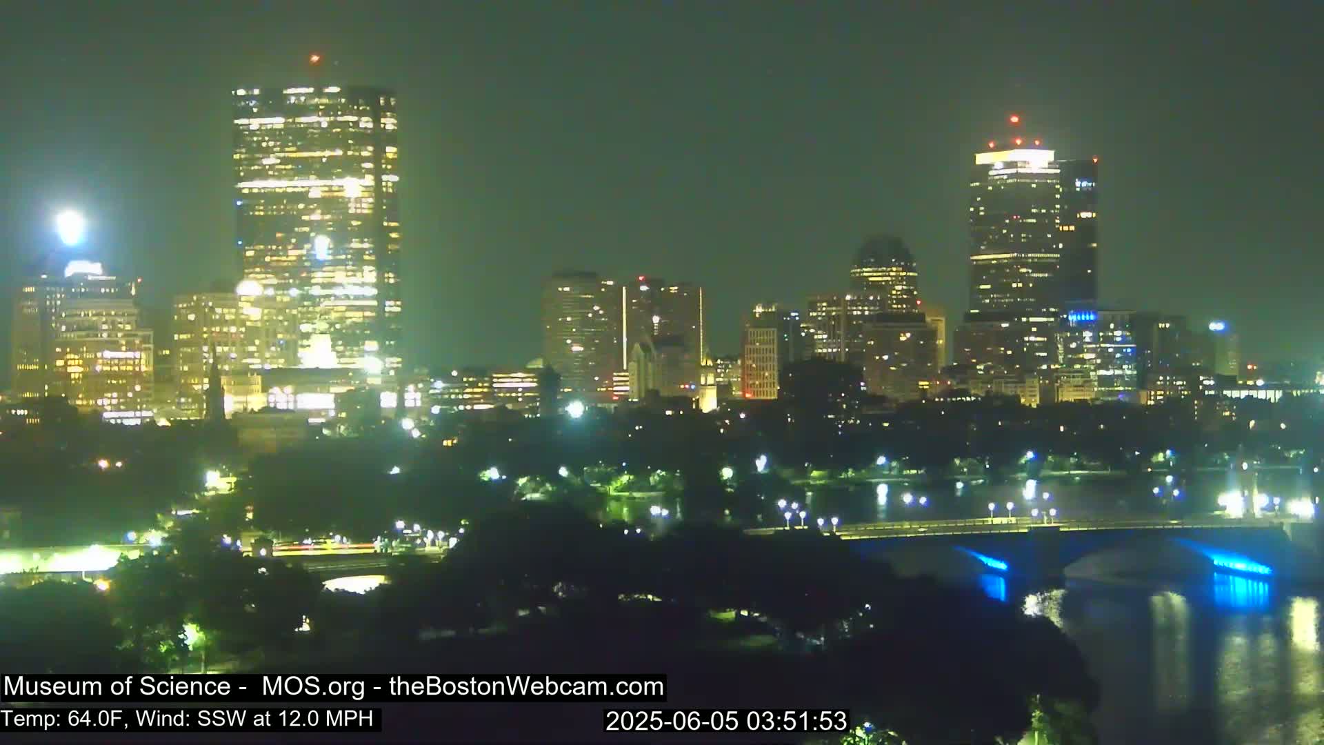 A nighttime cityscape with illuminated skyscrapers and a bridge over a body of water under a clear, dark sky with a south-southwesterly wind at 12 mph and a temperature of 64°F.