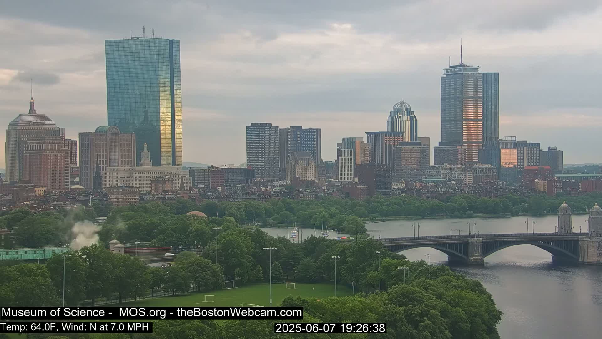 A city skyline with several tall buildings is seen across a river with a bridge, under an overcast sky.