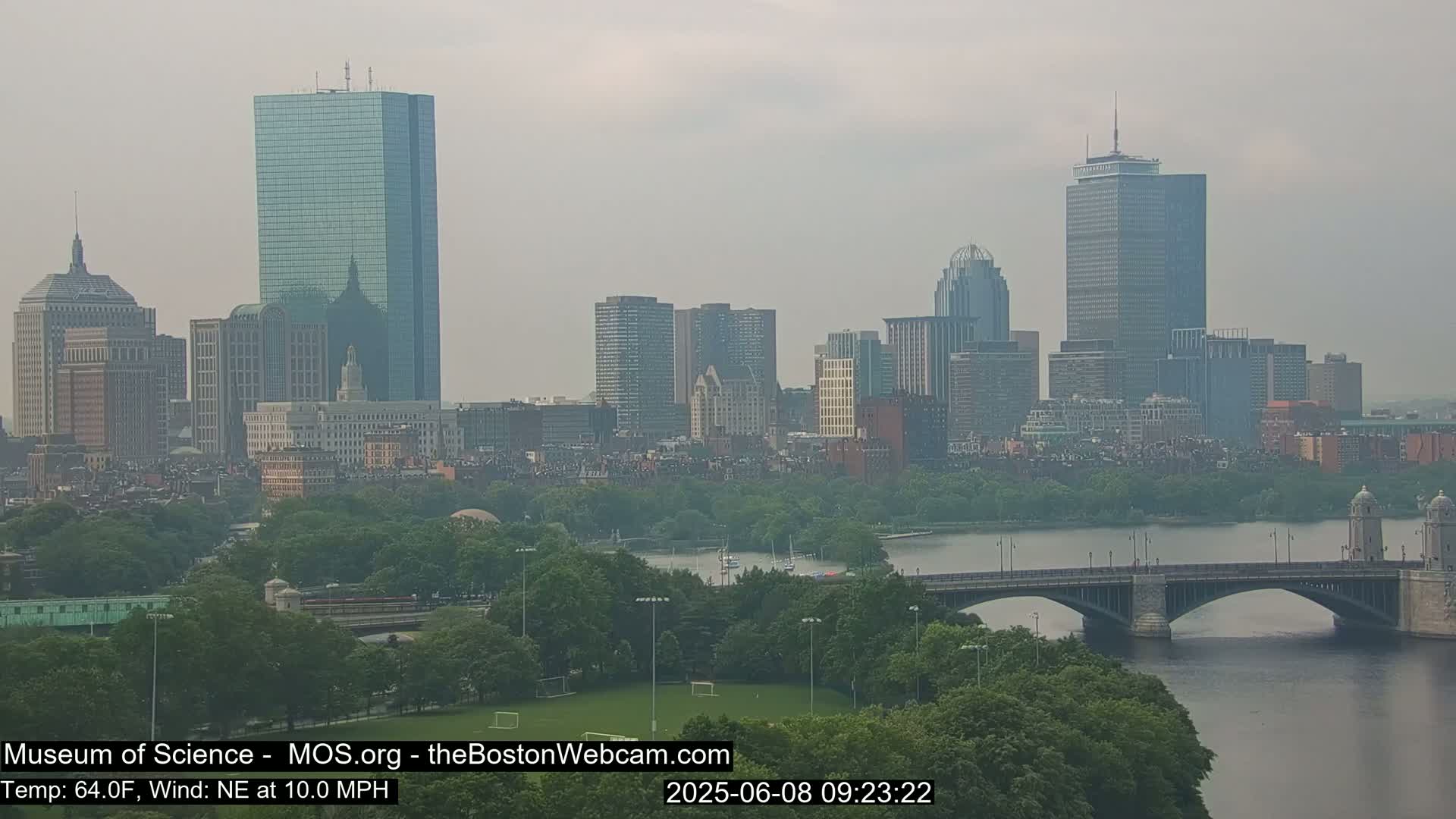 A hazy view of the Boston skyline across a river, with a bridge and green spaces in the foreground.