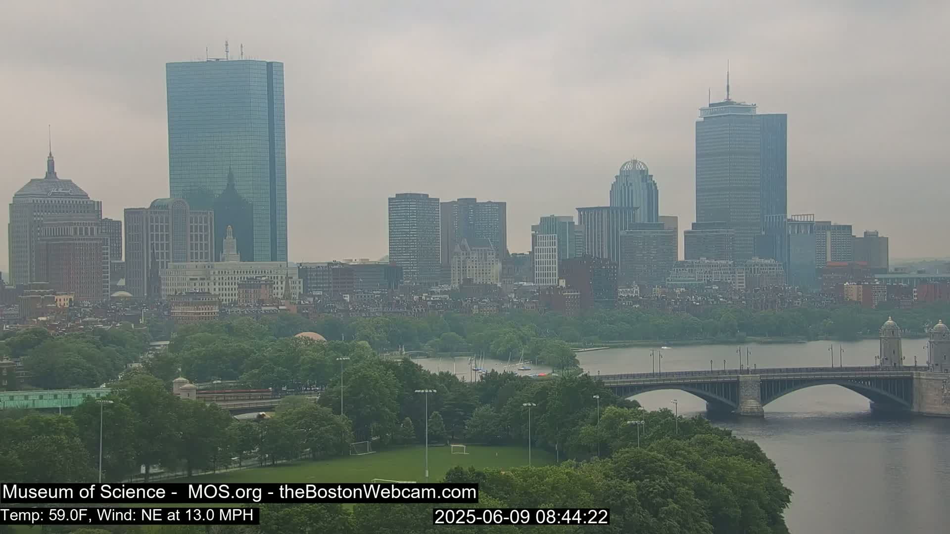 A hazy view of the Boston skyline, including several skyscrapers and a bridge spanning a river, on a day with a northeast wind of 13 mph and a temperature of 59°F.