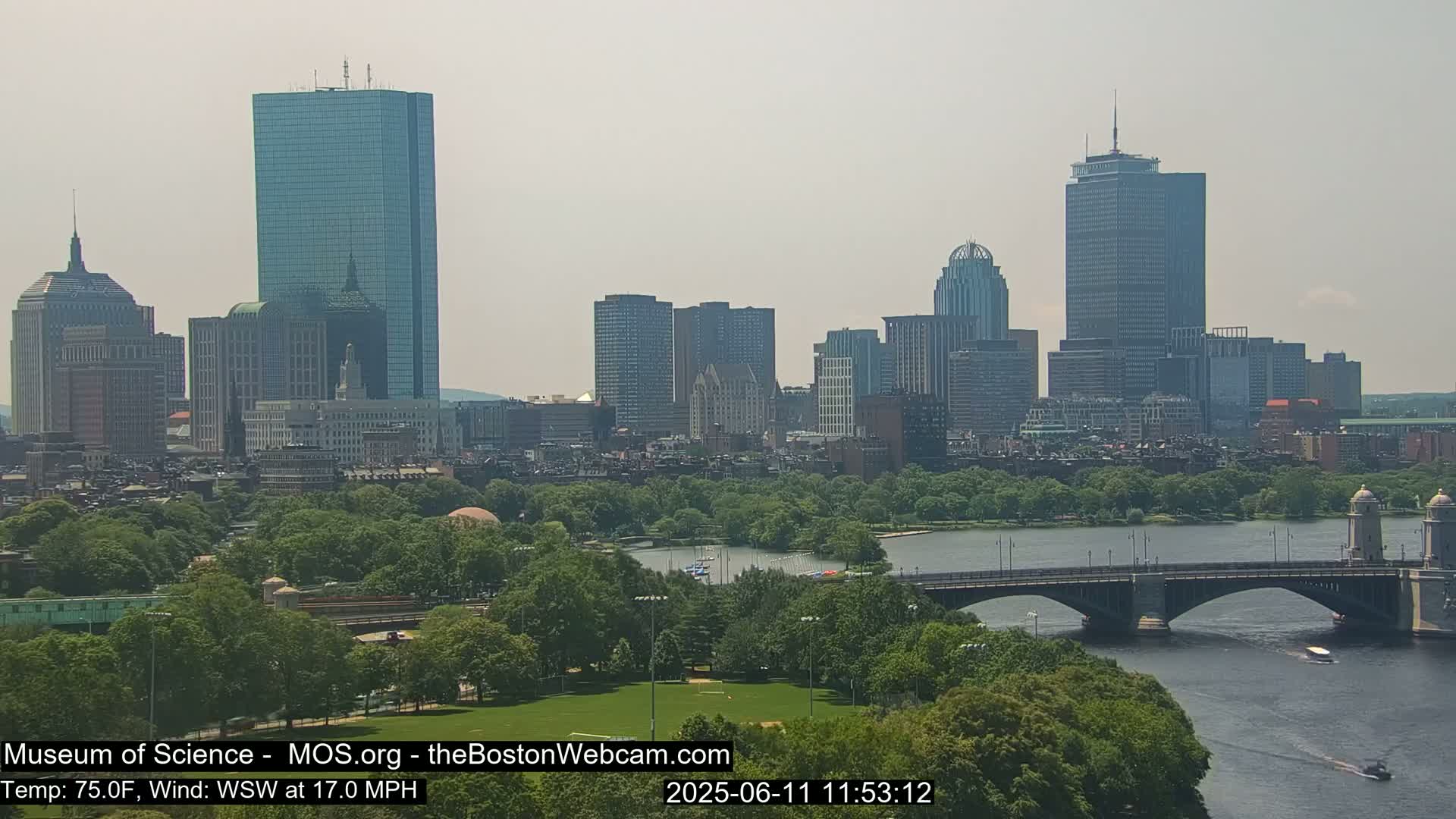 A hazy view of the Boston skyline across a river and park, with a bridge in the foreground.