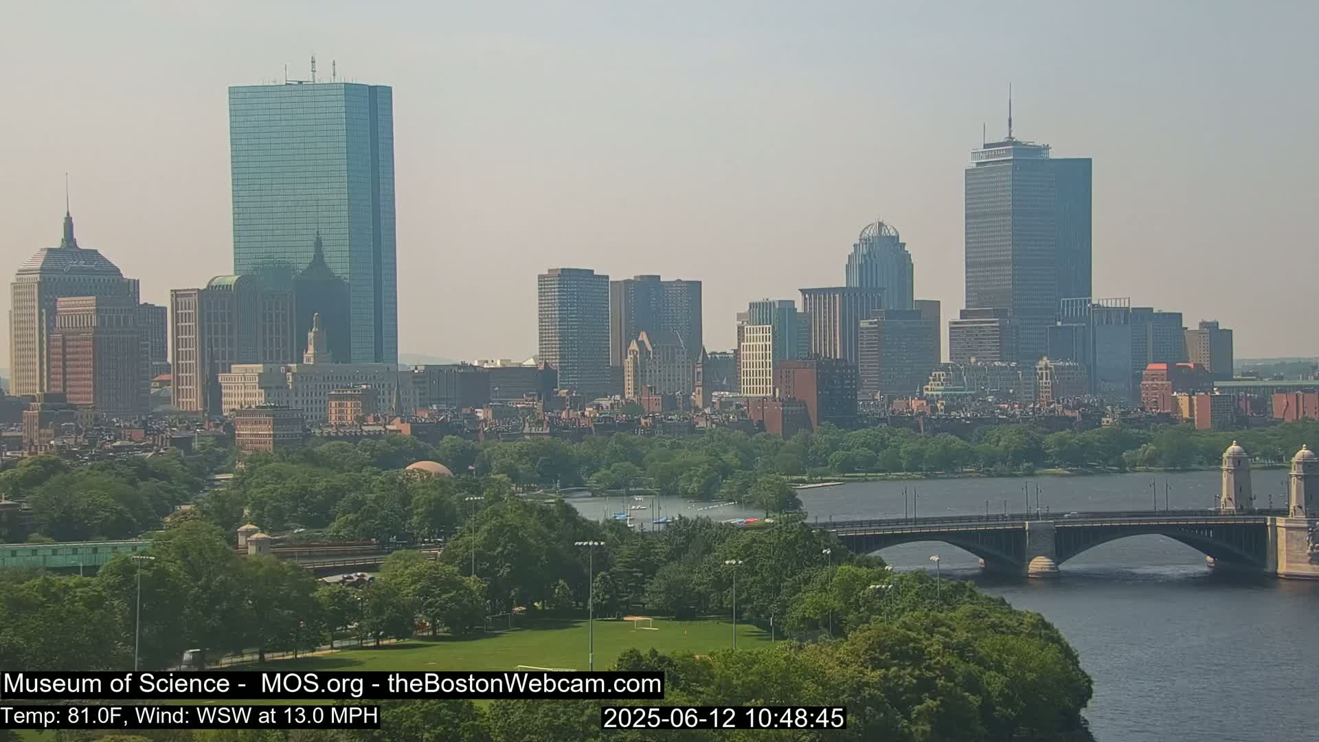 A hazy view of the Boston skyline across a river, with a bridge in the foreground and lush green trees lining the riverbanks under a partly sunny sky.
