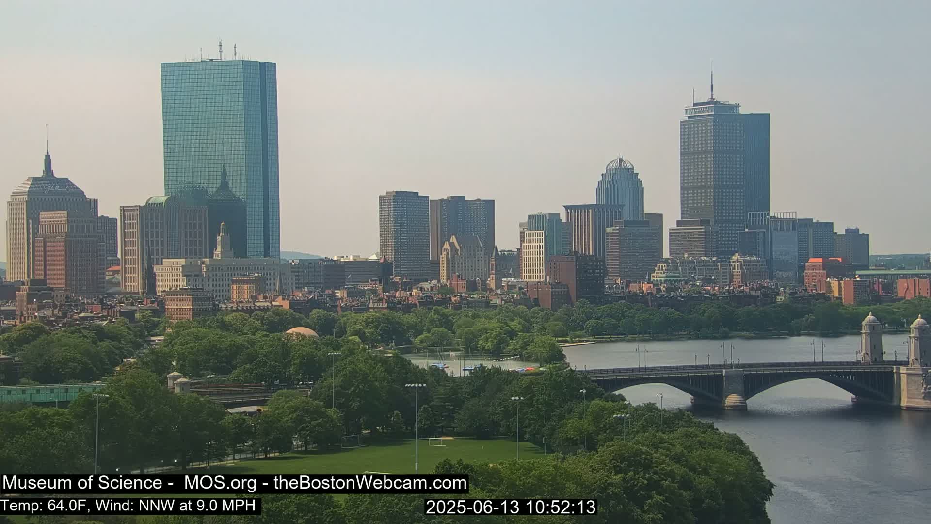 A cityscape with numerous skyscrapers and a bridge over a river is visible on a partly sunny day with a north-northwest wind at 9 miles per hour and a temperature of 64 degrees Fahrenheit.