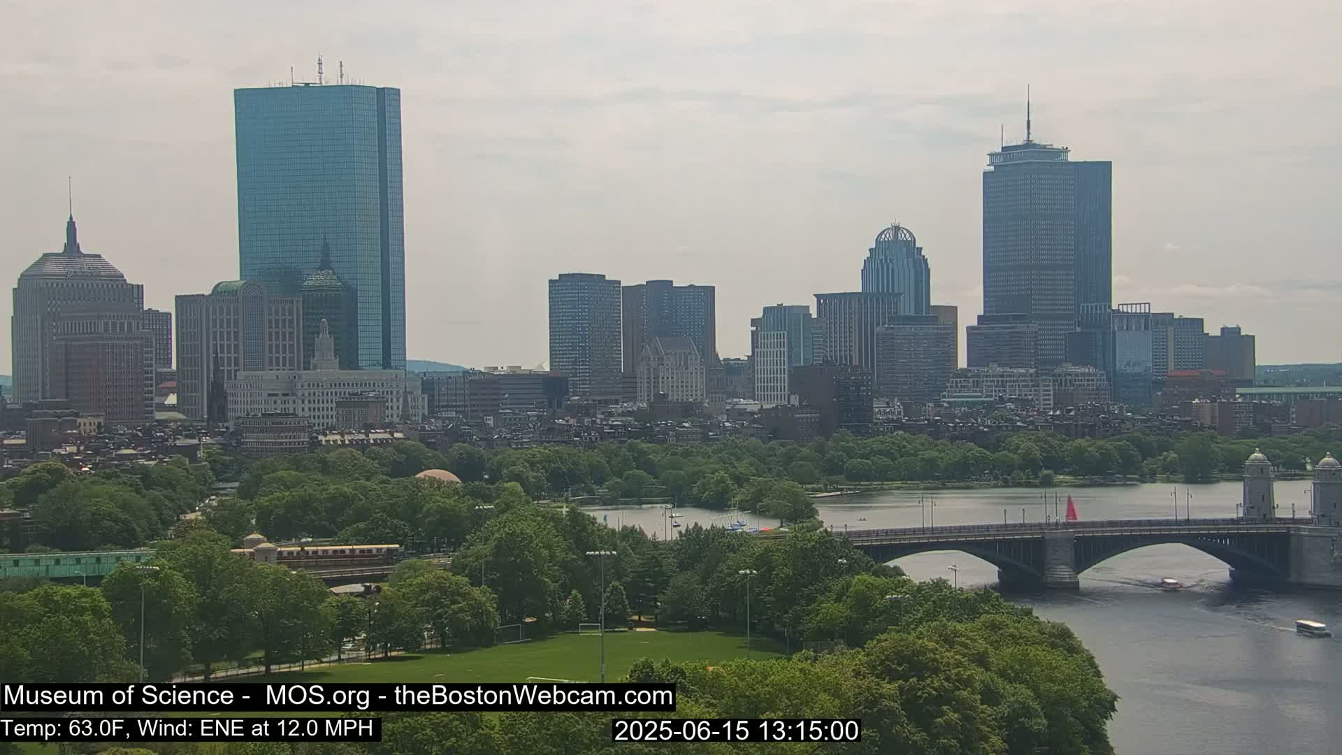 A hazy view of the Boston skyline, including several skyscrapers and a bridge spanning a river with lush green trees along the riverbanks, under a mostly cloudy sky.