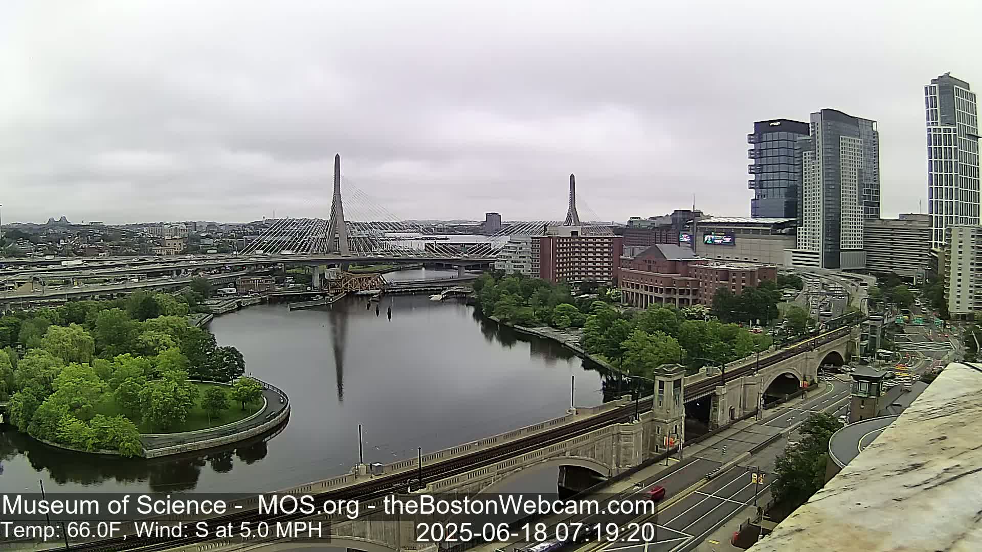 A partly cloudy view of a city with two cable-stayed bridges over a river, surrounded by green trees and buildings, under a mostly overcast sky.