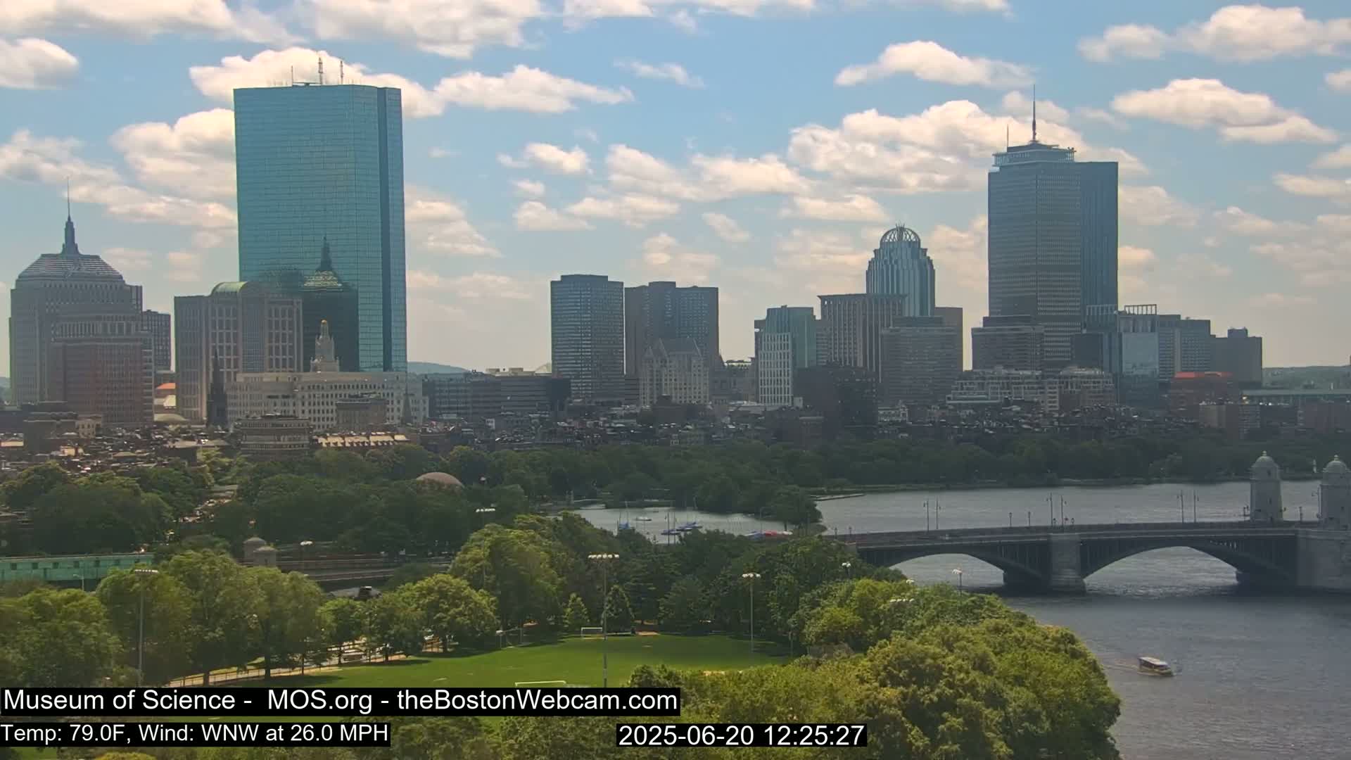 A partly cloudy day shows a cityscape across a river with a bridge, featuring numerous skyscrapers and lush green spaces.