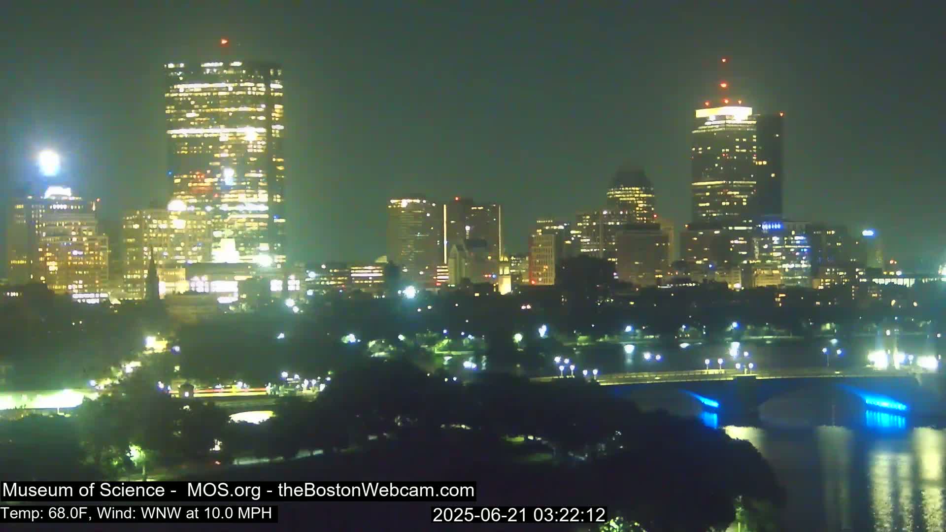 A nighttime view of a city skyline across a body of water, with a bridge illuminated in blue under a clear sky and a temperature of 68°F and a wind of 10 mph from the west-northwest.