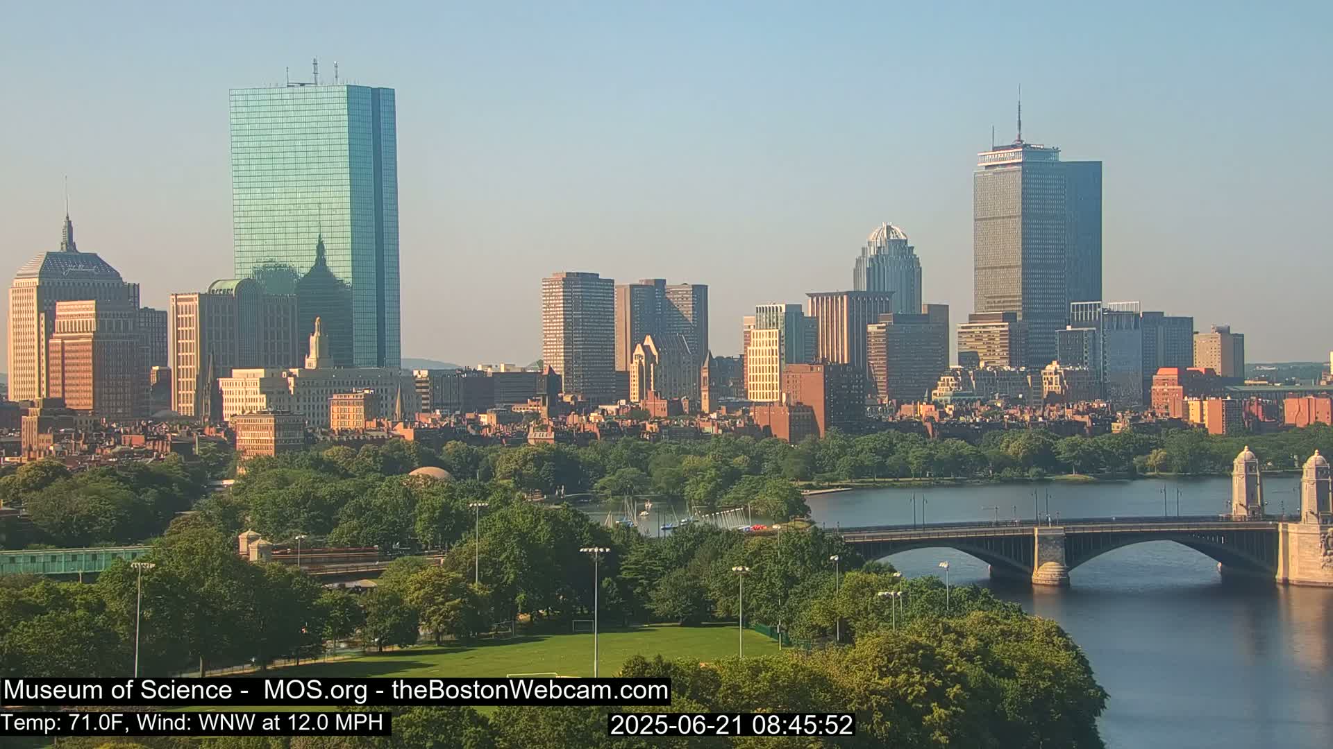 A city skyline with numerous skyscrapers and other buildings is visible across a calm river, with a bridge in the foreground and lush green trees lining the riverbanks on a clear, sunny day with a temperature of 71°F and a west-northwest wind at 12 mph.