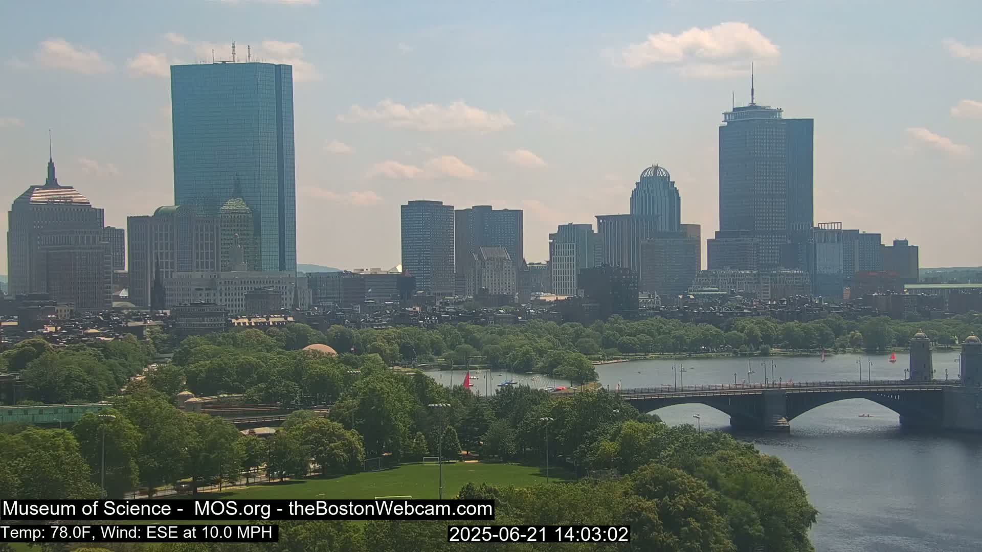A city skyline with numerous skyscrapers is visible across a body of water, with a bridge spanning the river and lush green trees lining its banks, under a partly cloudy sky.