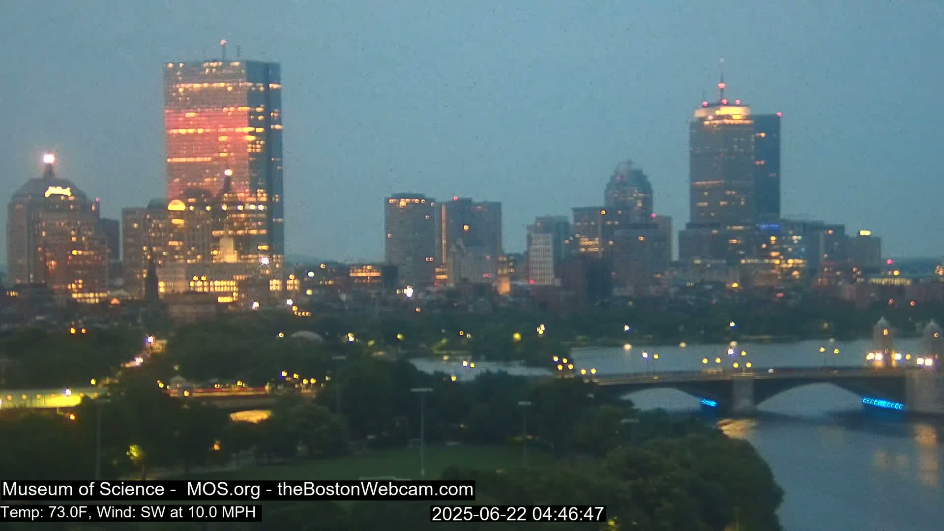 A nighttime view of a city skyline illuminated by lights, with a bridge spanning a river in the foreground, under a hazy, dusky sky.