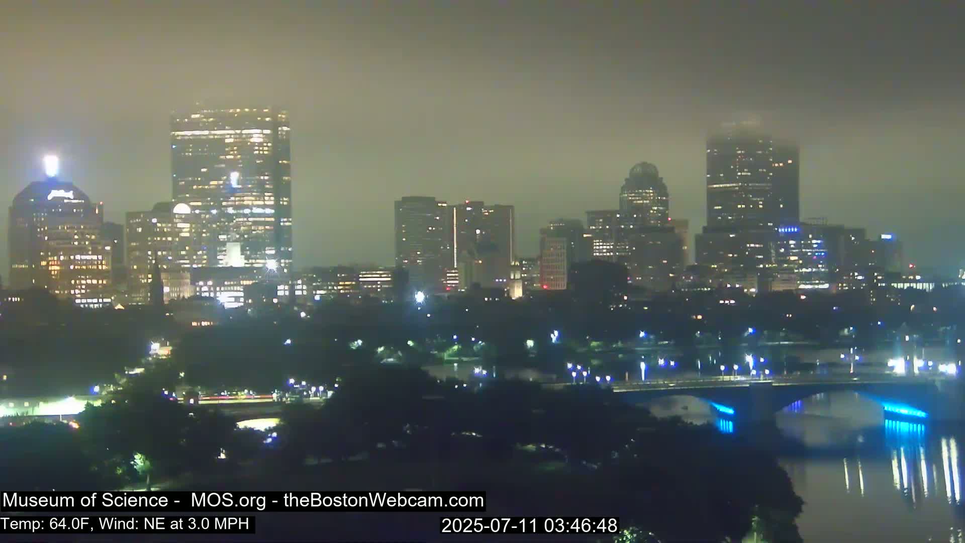A nighttime view of a city skyline shrouded in fog, with a bridge and river in the foreground.