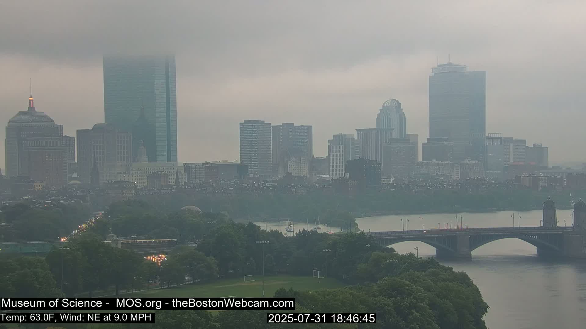 A hazy cityscape with tall buildings and a bridge over a river is visible on a cloudy day with a northeast wind.