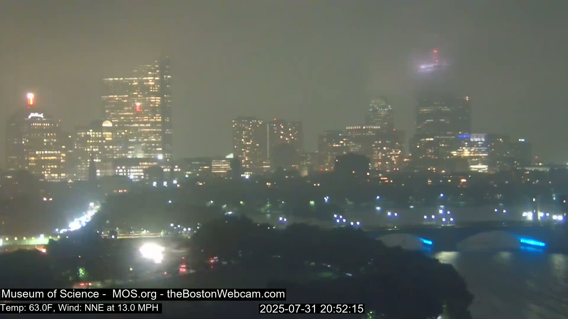 A hazy, nighttime view of a city skyline with illuminated buildings and a bridge across a body of water, under a NNE wind of 13.0 MPH and a temperature of 63.0°F.