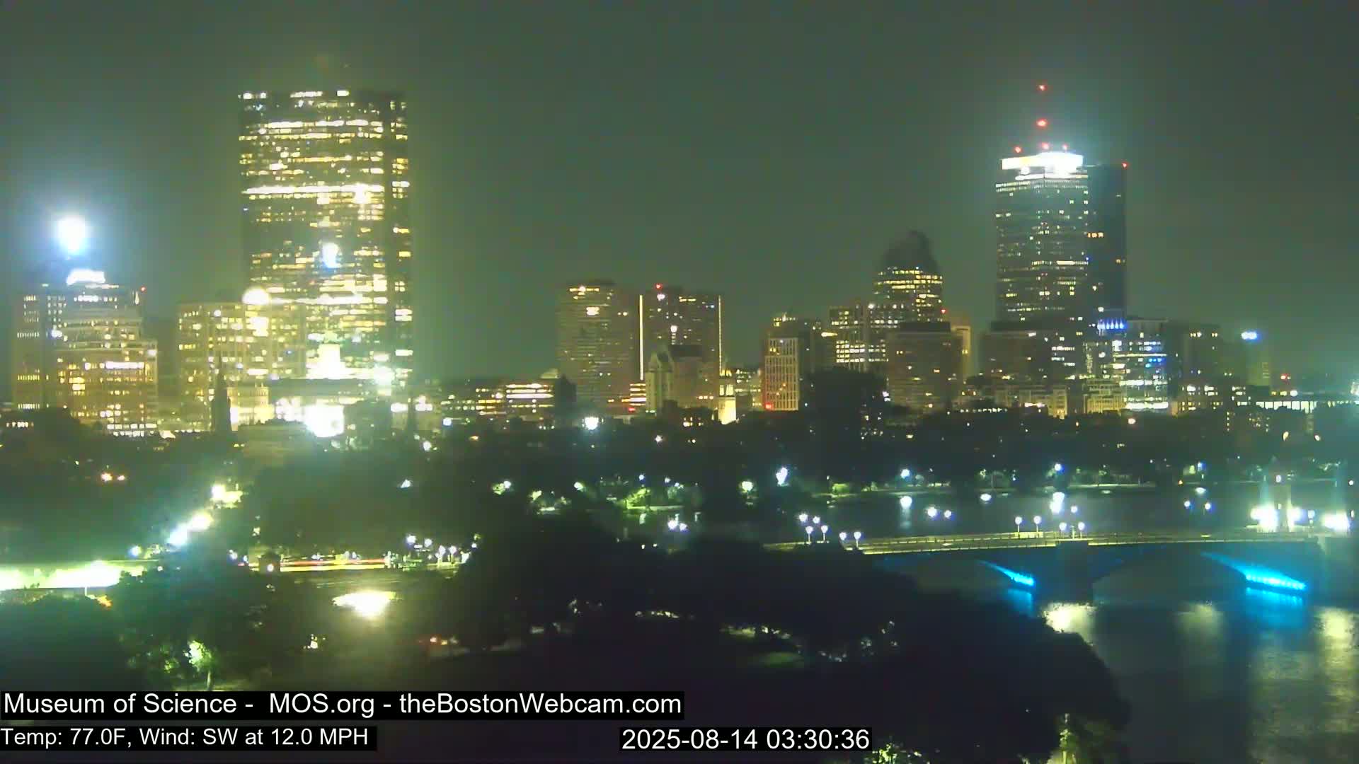 A nighttime cityscape with brightly lit skyscrapers and a bridge over a body of water, under a clear sky with a southwest wind at 12 mph and a temperature of 77°F.