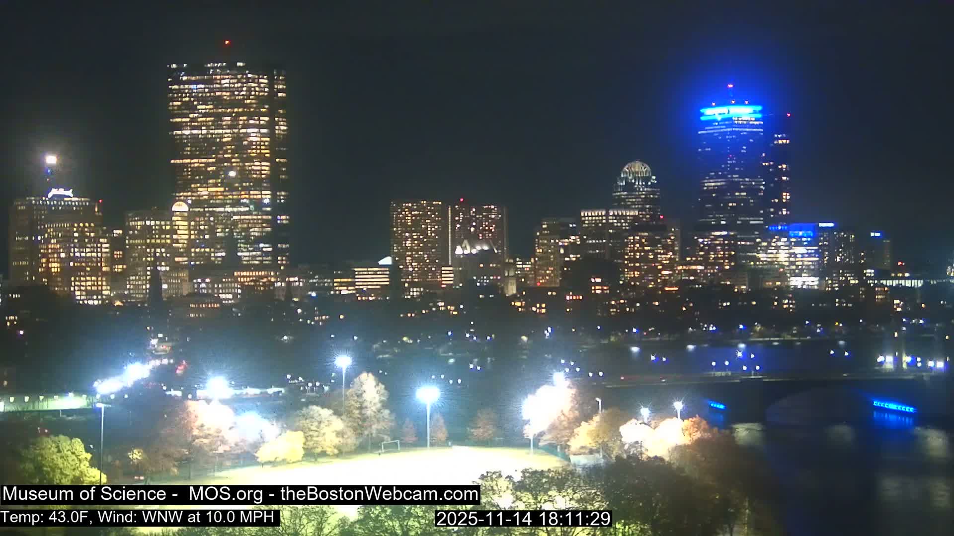 A clear night view of a city skyline features numerous illuminated buildings, including one with a bright blue top, overlooking a lit park with autumn trees and a bridge spanning a river, under conditions of 43.0F with a WNW wind at 10.0 MPH.