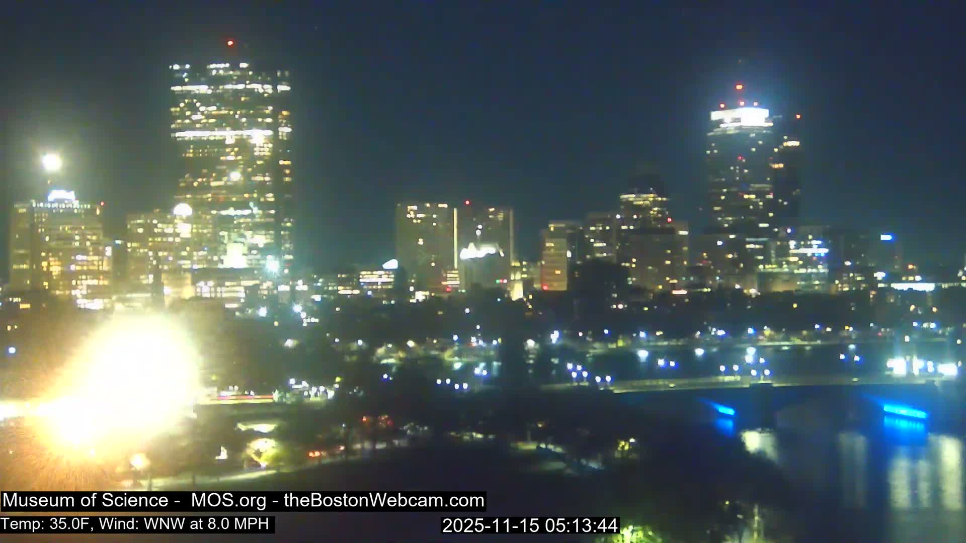 A clear, dark night in the city reveals a skyline of brightly lit buildings and a bridge with blue lights reflecting on a body of water, accompanied by a strong light flare in the lower left, with a temperature of 35.0F and a WNW wind at 8.0 MPH.