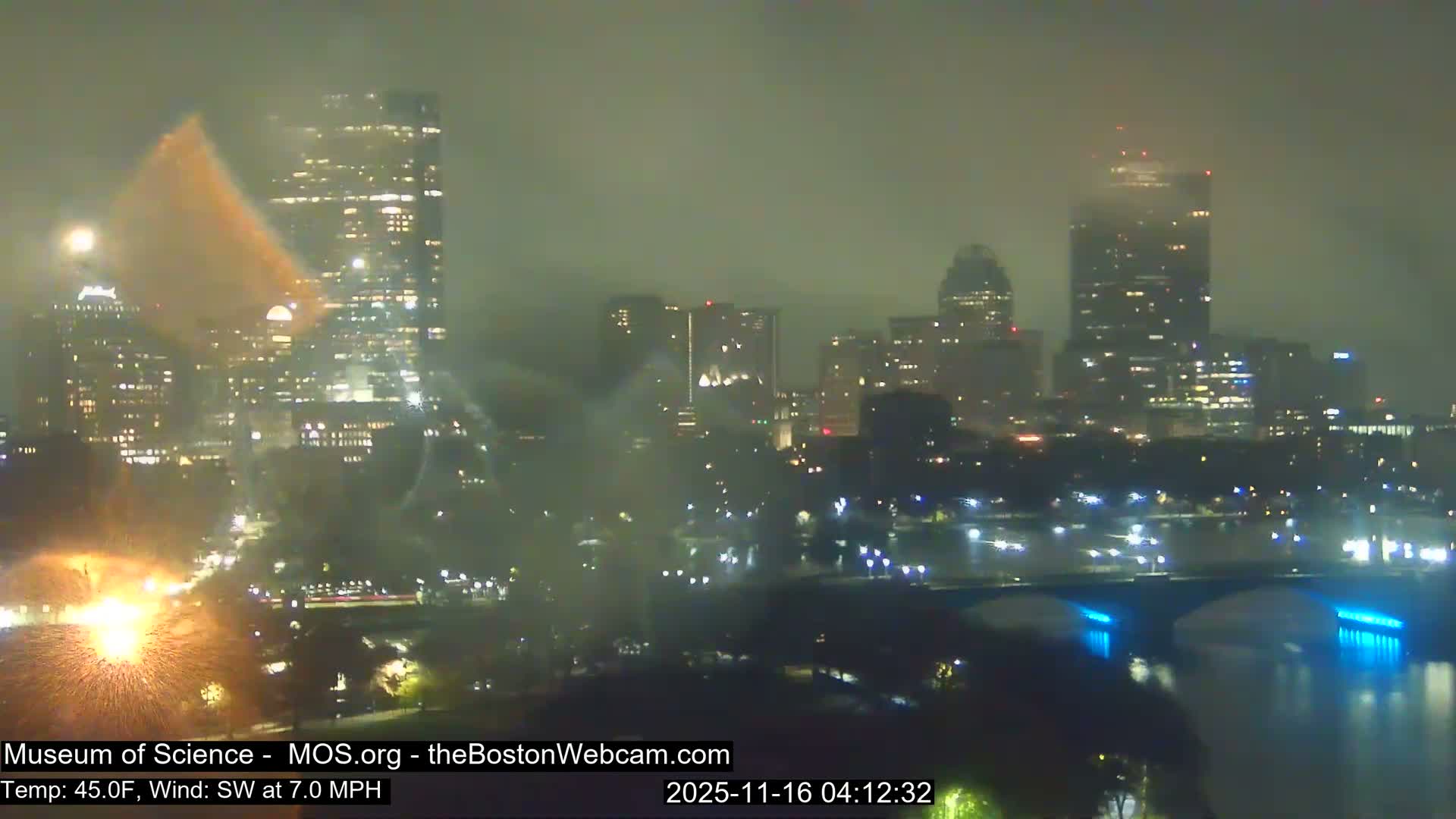 A foggy nighttime cityscape is illuminated by numerous buildings and a brightly lit bridge over a river, with a prominent burst of orange sparks resembling fireworks on the lower left.