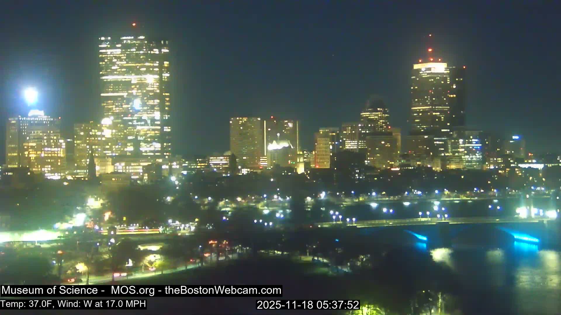 A foggy nighttime cityscape is illuminated by numerous buildings and a brightly lit bridge over a river, with a prominent burst of orange sparks resembling fireworks on the lower left.