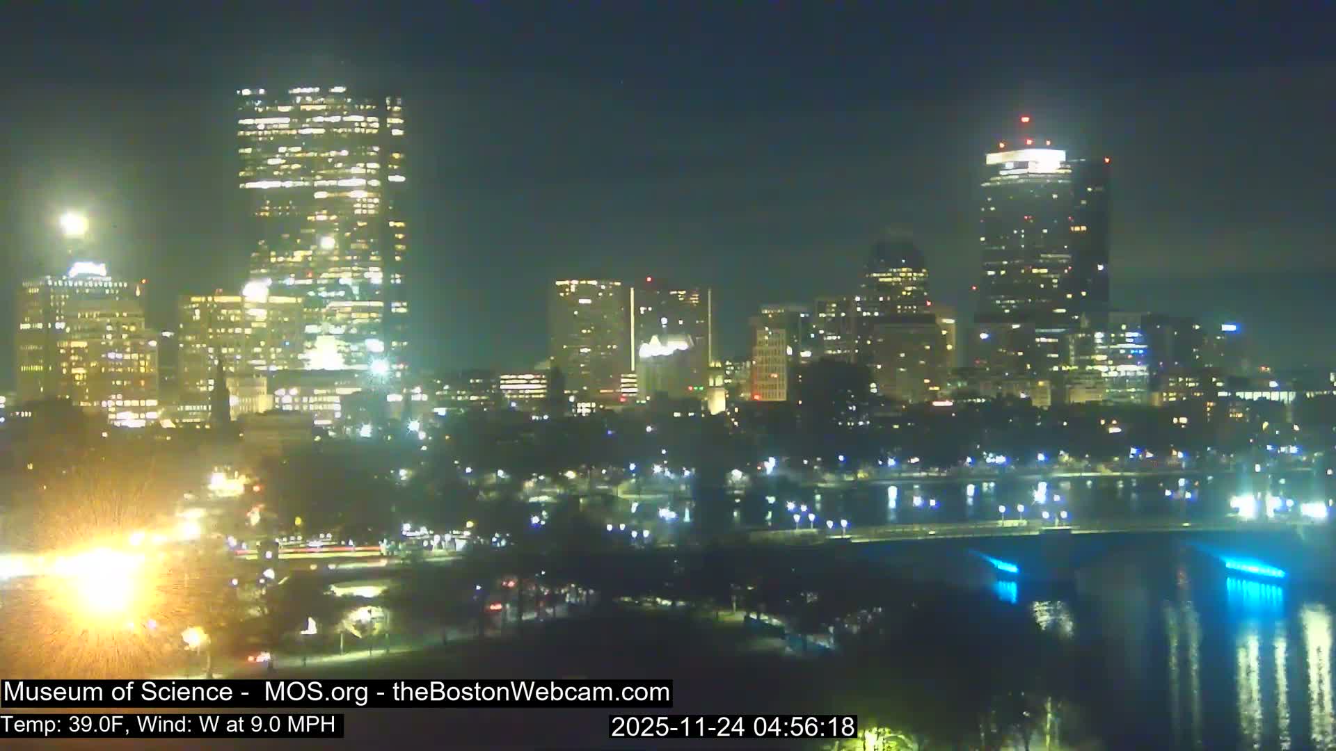 A nighttime view of a brightly lit city skyline features numerous illuminated skyscrapers and buildings, some reflecting on a body of water with a bridge adorned with blue lights, all under clear skies with a temperature of 39.0F and a west wind at 9.0 MPH.