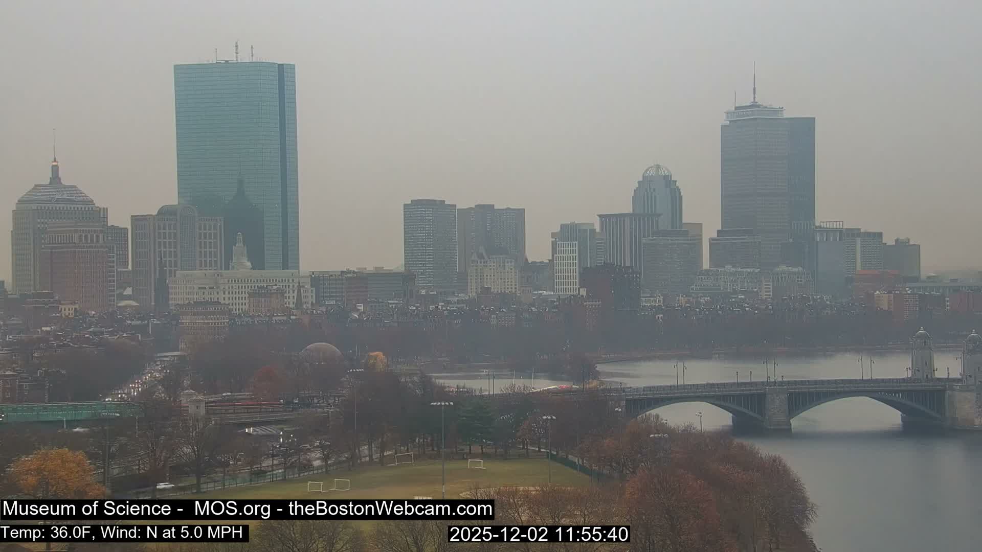 A hazy and overcast daytime view overlooks a bustling city skyline featuring numerous tall buildings, a wide river with several arch bridges, and foreground parks with leafless trees and active roads.