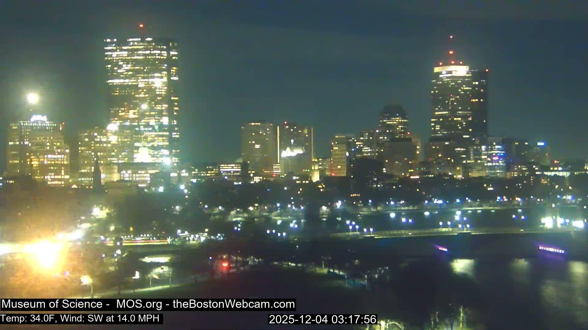 A sprawling city skyline glows brightly at night with countless lights from buildings, reflected on a body of water, under a clear, dark sky.