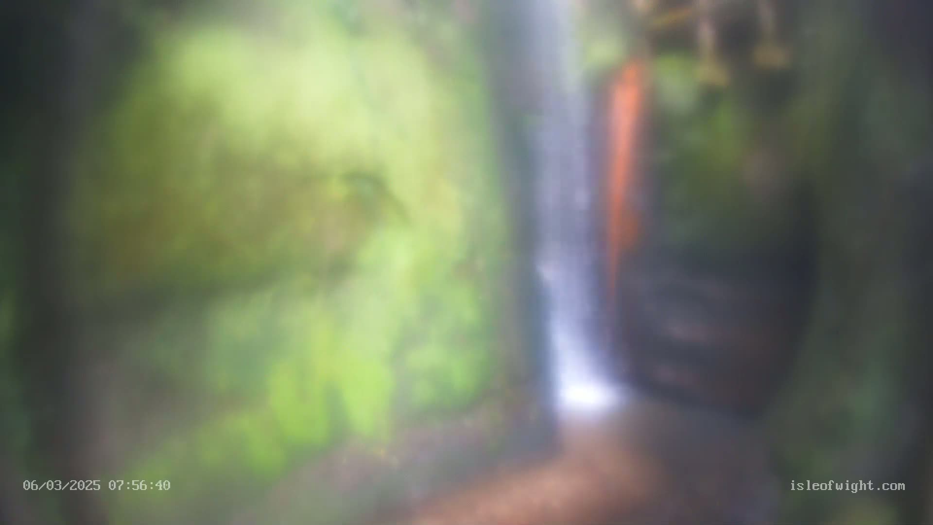 A blurry image shows a waterfall cascading down a mossy rock face in a humid environment.