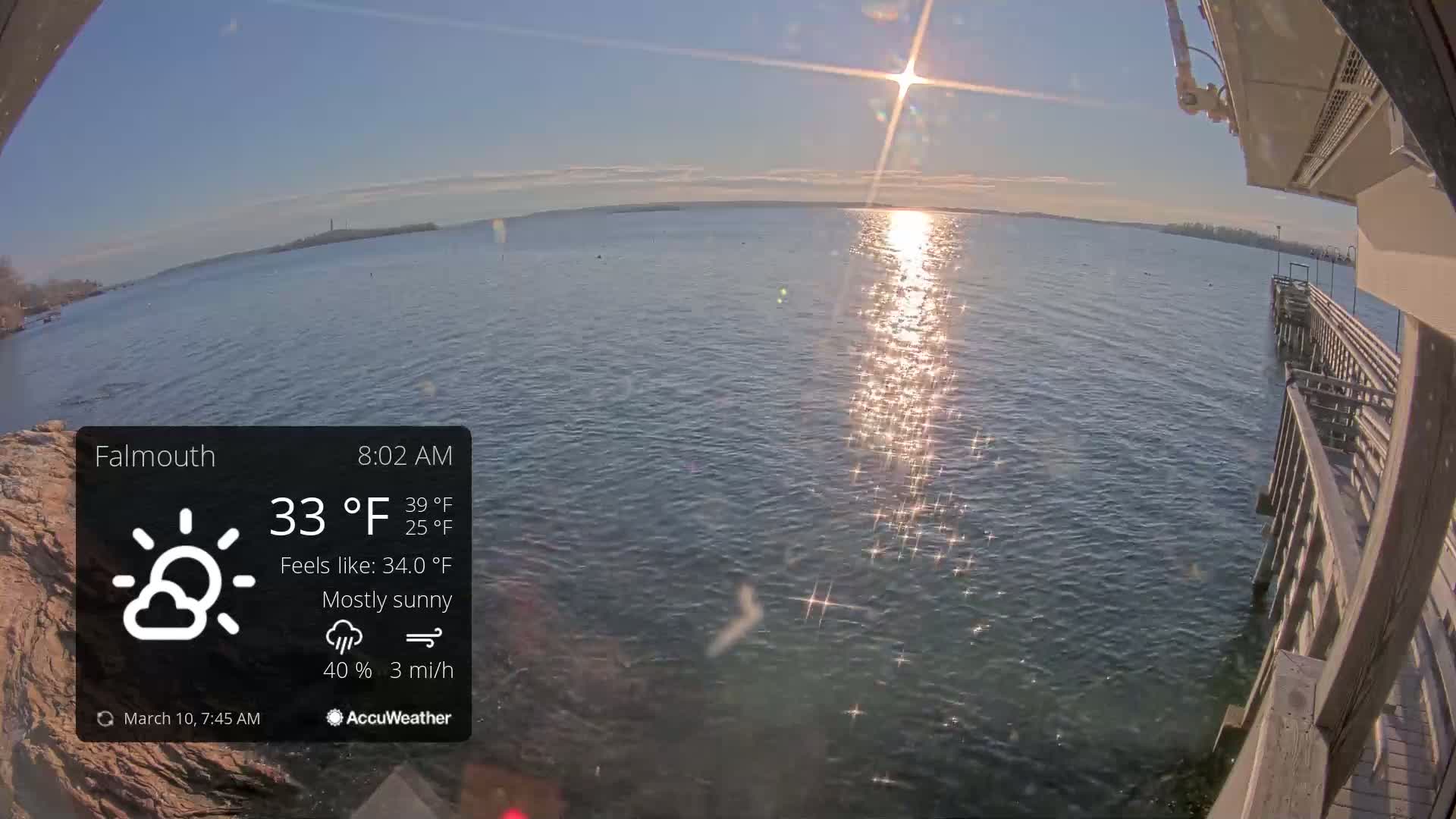 A mostly sunny morning scene shows a calm body of water reflecting sunlight near a wooden pier, with a distant landmass visible on the horizon.