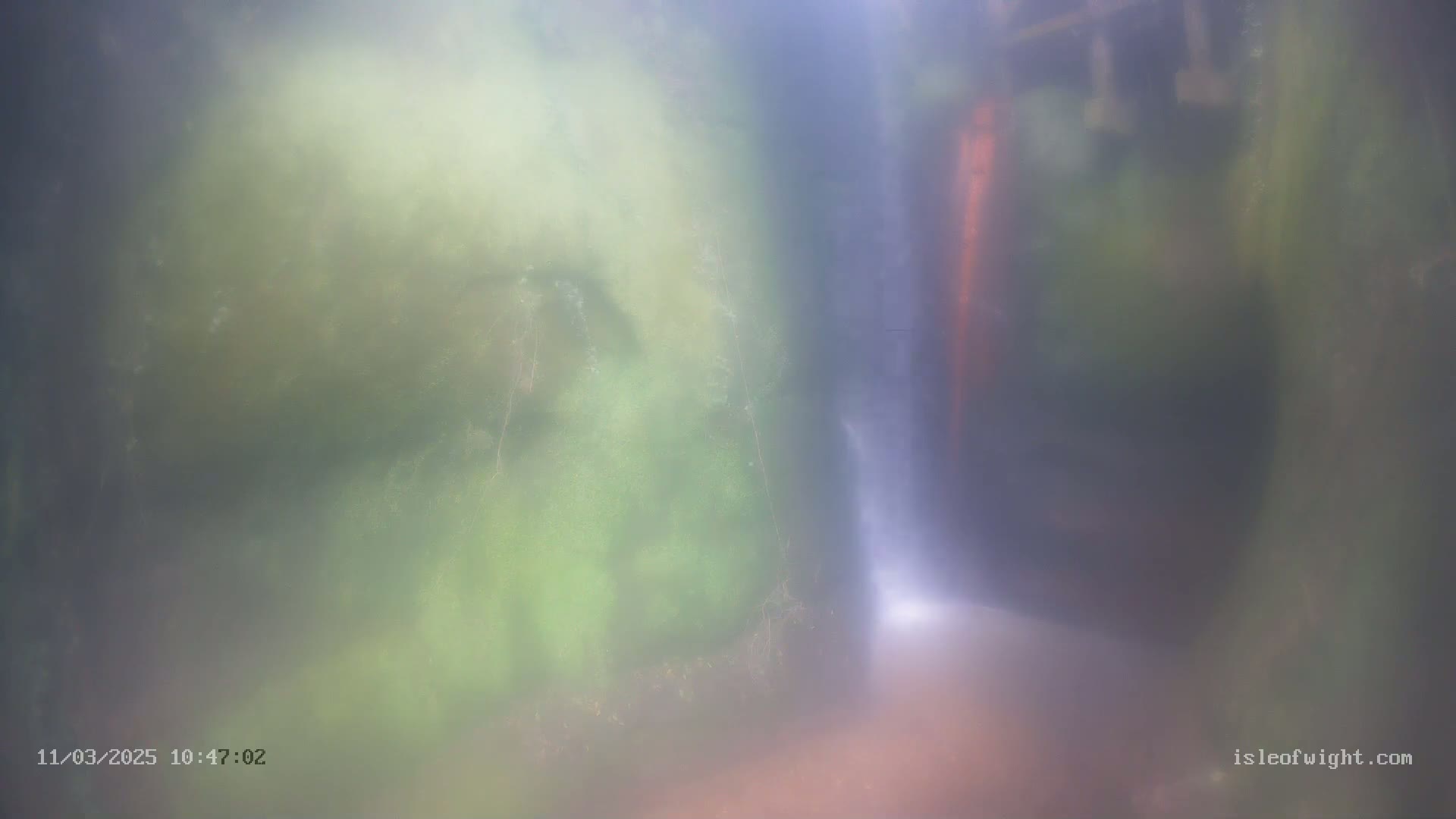A blurry underwater view shows a partially visible waterfall flowing between mossy rock formations in hazy water.