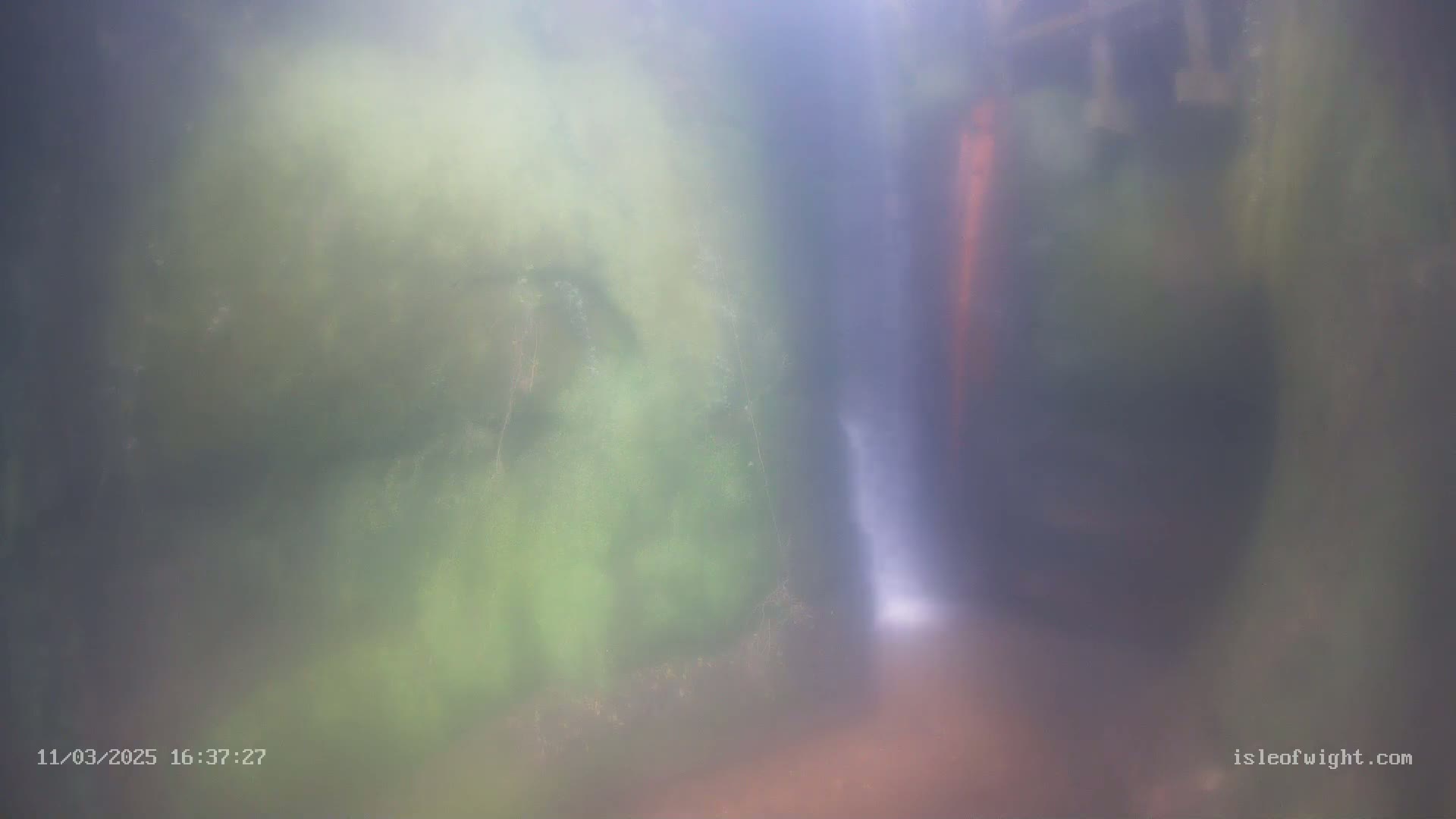 A blurry underwater view shows a small waterfall cascading between mossy green rocks in hazy water.