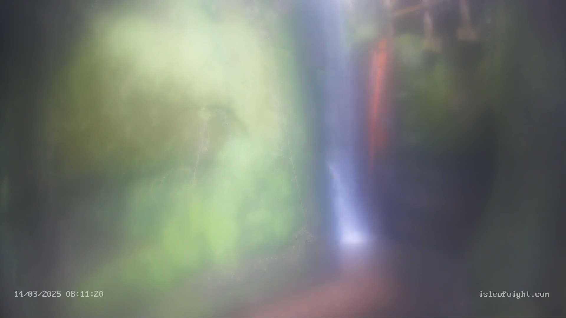 A blurry image shows a waterfall partially obscured by mist or fog in a lush green environment.
