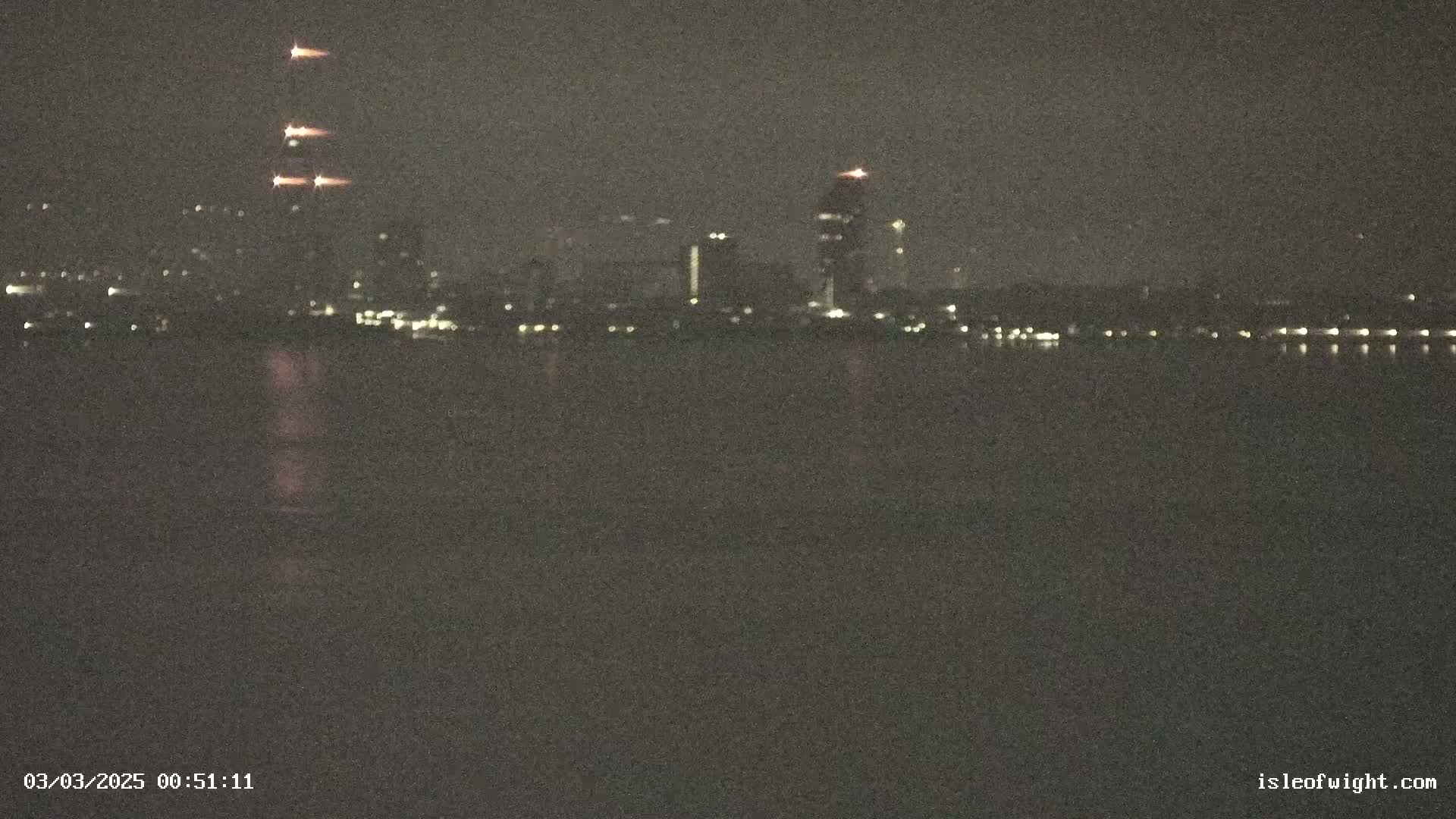 A nighttime view across dark water shows a distant city skyline with several tall buildings illuminated, one of which has bright lights ascending vertically.