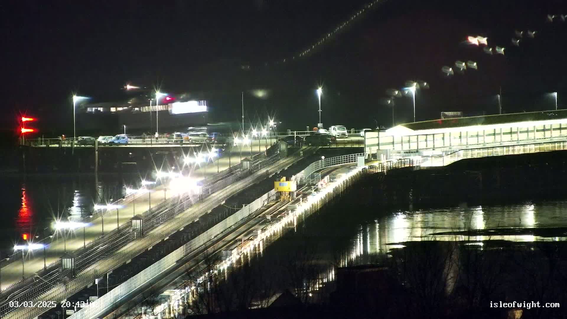 A ferry terminal at night is brightly lit, with a vehicle waiting on the tracks to board a ferry, and calm water reflecting the lights.