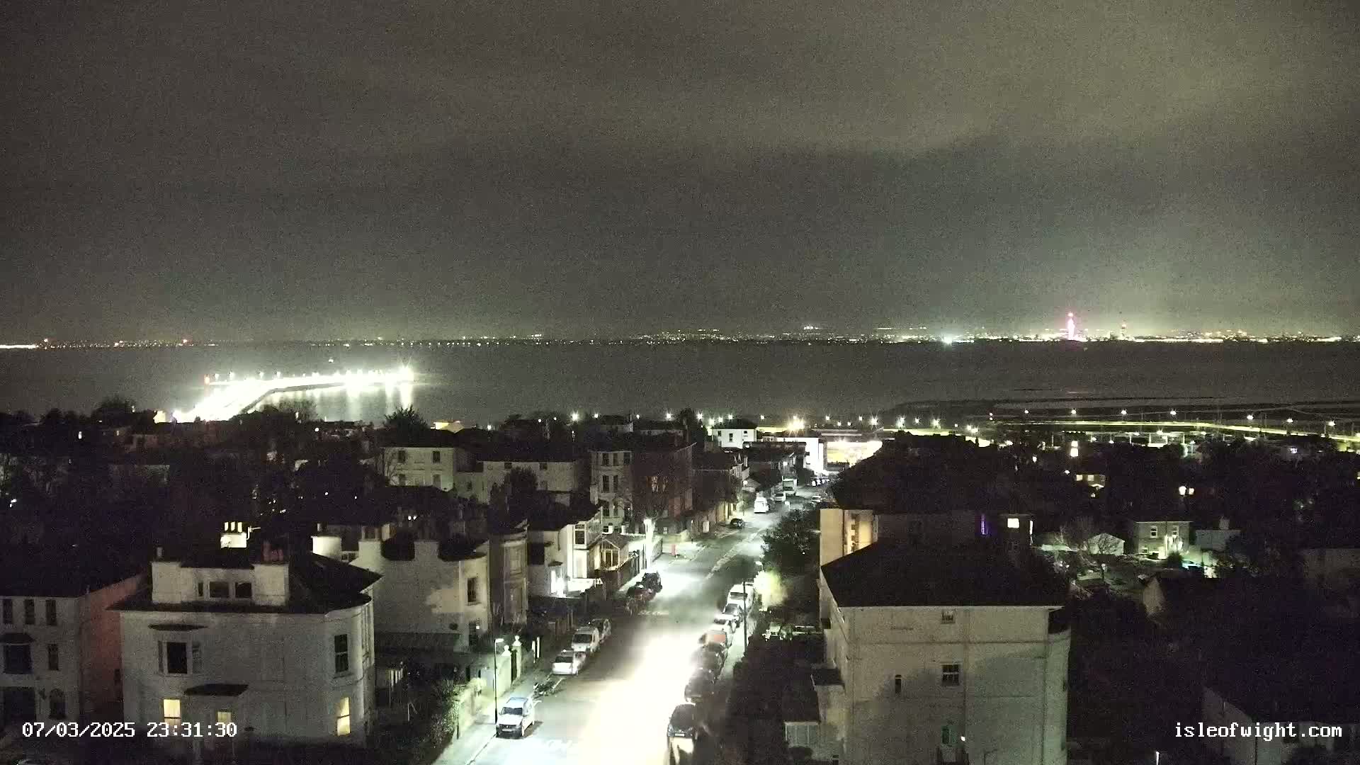 A nighttime, overcast view of a seaside town with a long pier extending into a dark body of water, illuminated by lights, and a city skyline visible in the distance.
