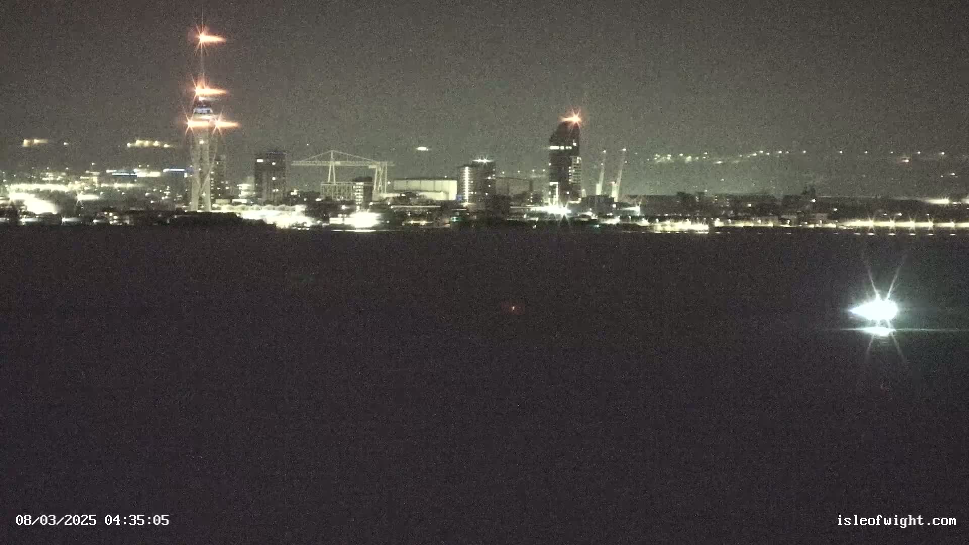 A nighttime view across dark water shows a city skyline with several tall buildings, lit-up structures, and cranes, under a clear, dark sky.
