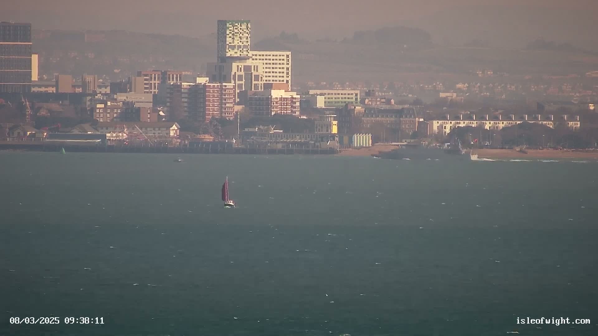 A sailboat is on a somewhat choppy sea in front of a hazy cityscape.
