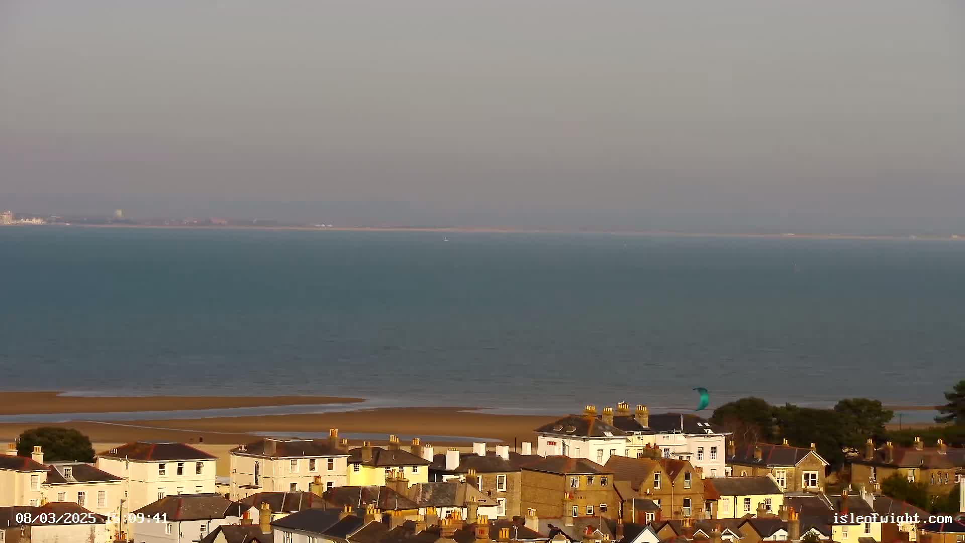 A kitesurfer glides over a calm, light-blue sea next to a sandy beach, overlooking a town of closely-packed, light-brown buildings.