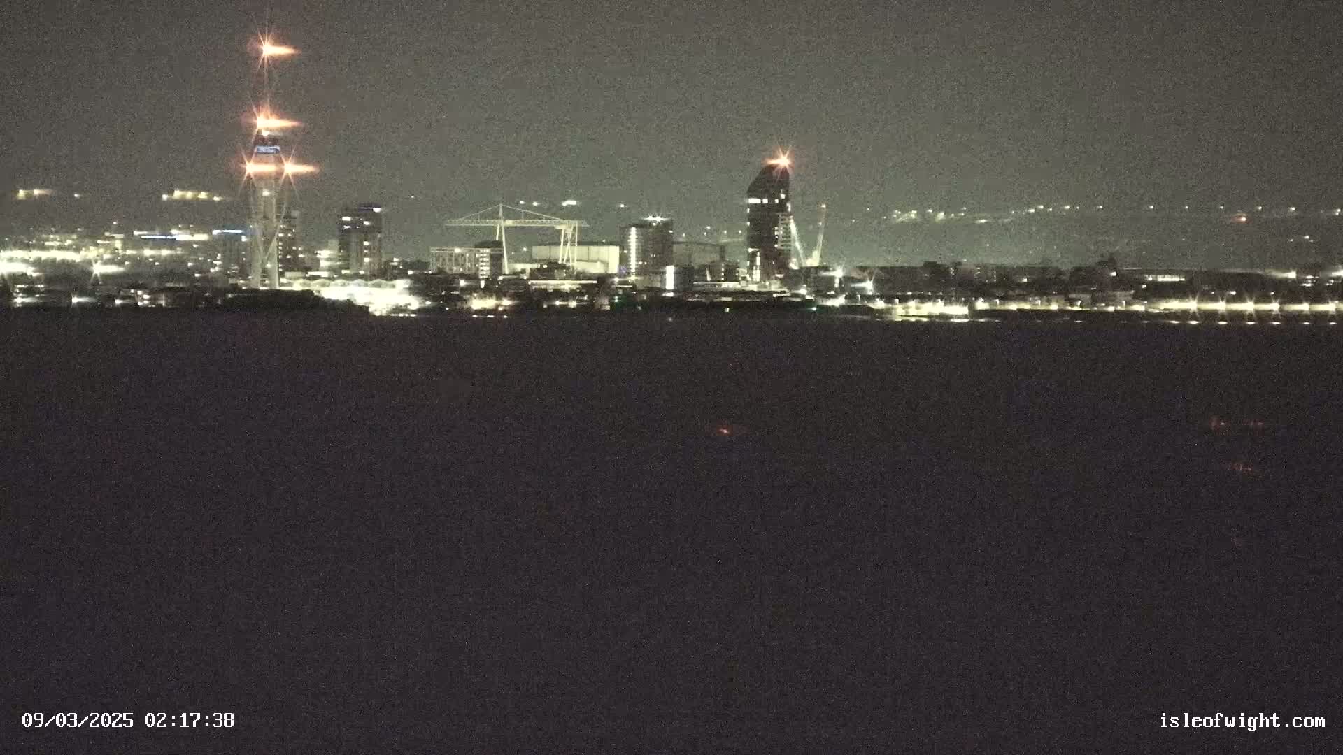A nighttime view of a city skyline across a body of dark water, illuminated by city lights.