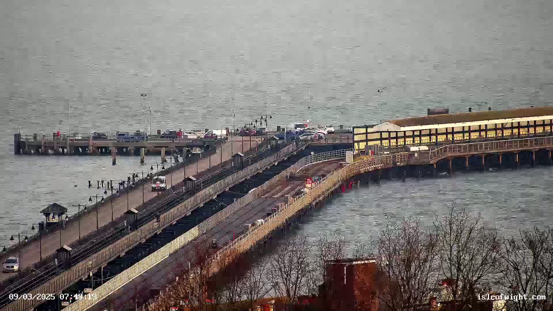 A long pier extends over a choppy, gray body of water, with vehicles parked at its end and a few cars traveling along its roadway.