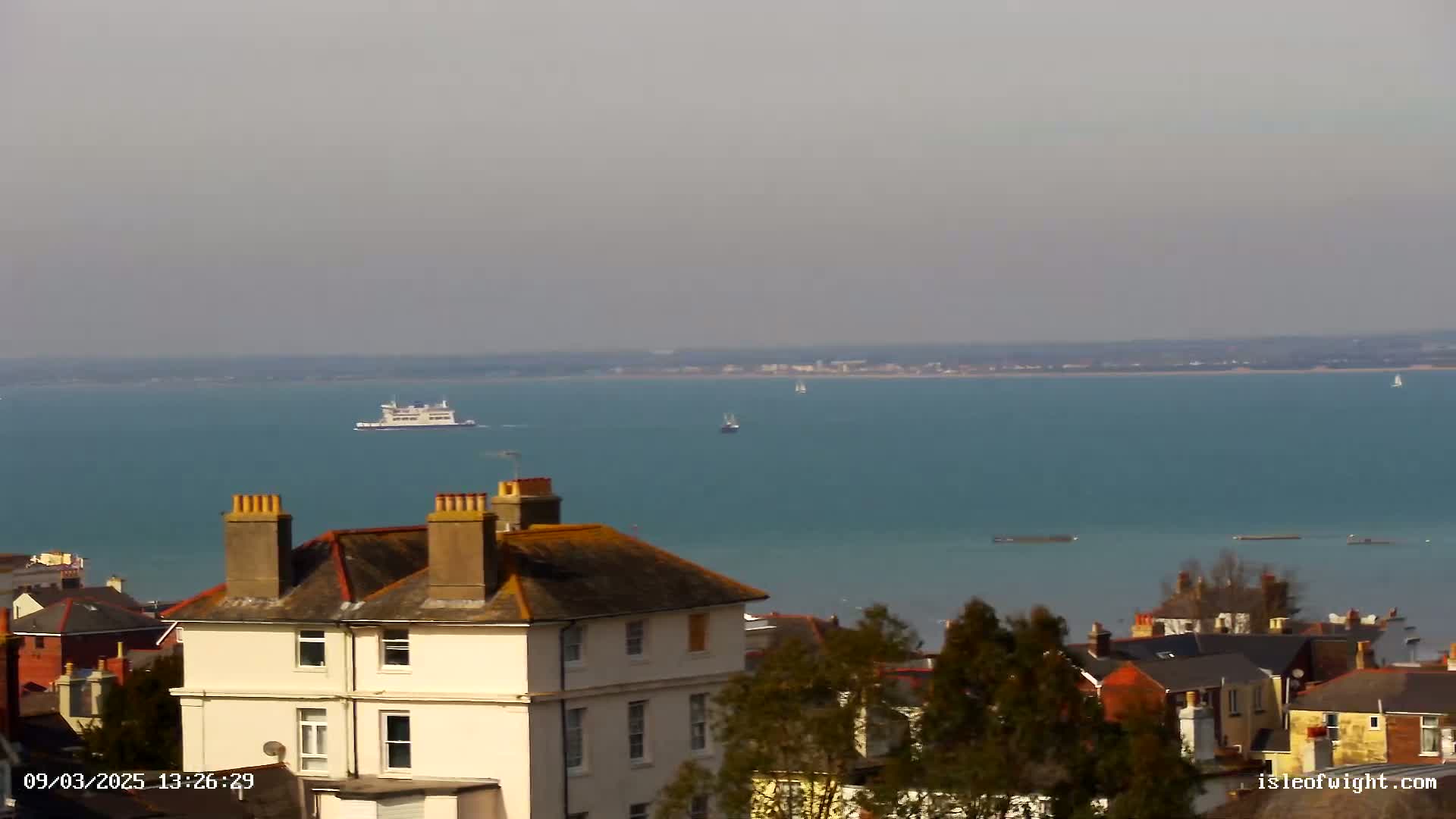 A white building with a gray roof sits atop a hill overlooking a calm teal sea where a large ferry and several smaller vessels are visible under a hazy sky.