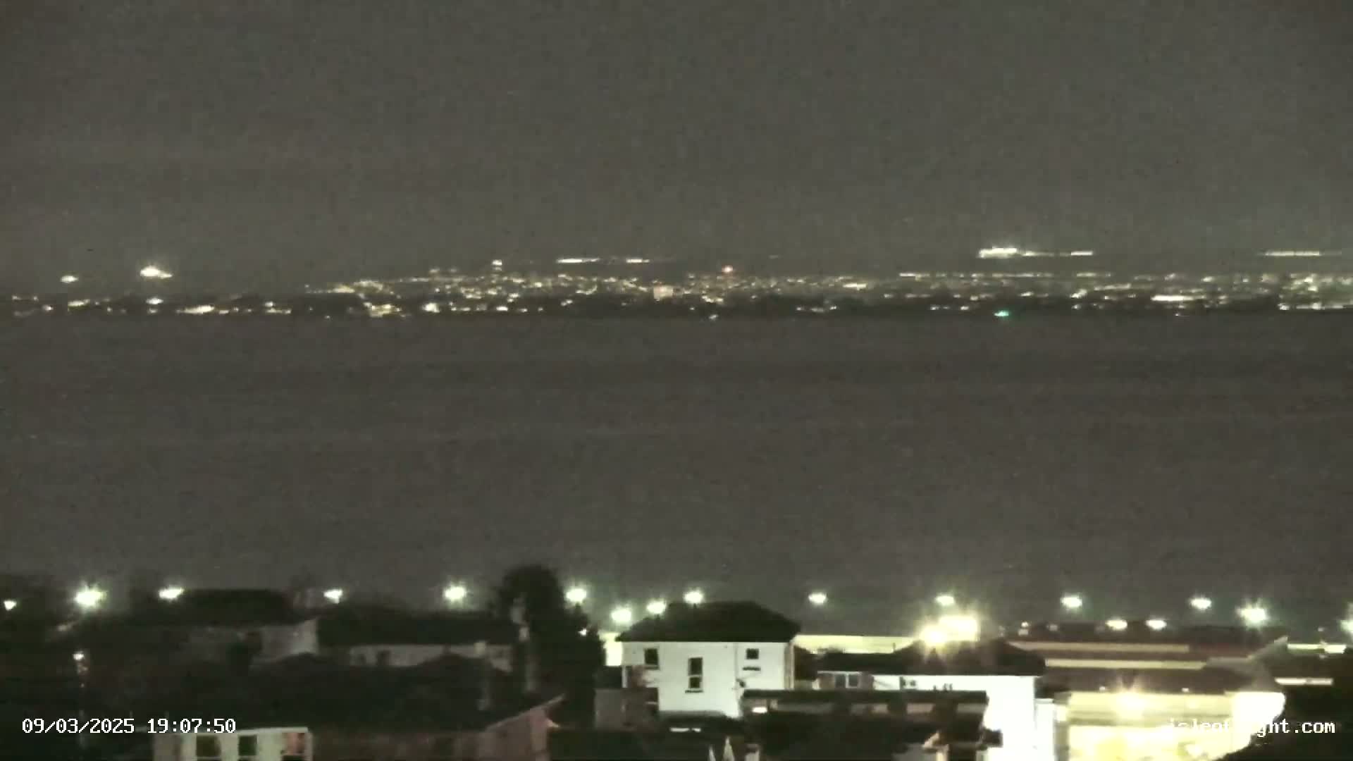A nighttime view shows a dark body of water with a distant city skyline faintly illuminated, and in the foreground, a small residential area with a few lit buildings.