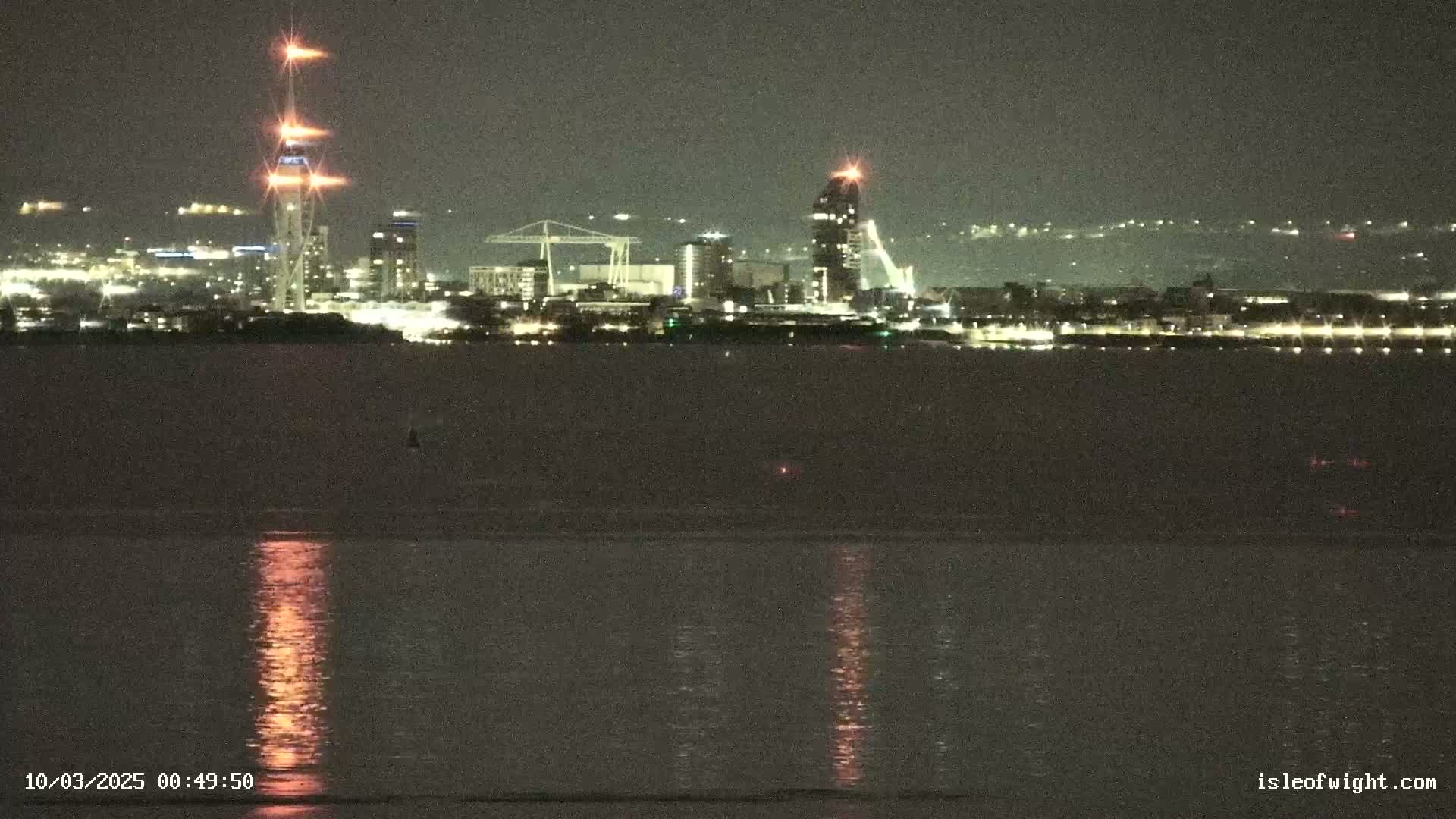 A nighttime view of a city skyline across a calm body of water, with several tall buildings illuminated against a dark sky.