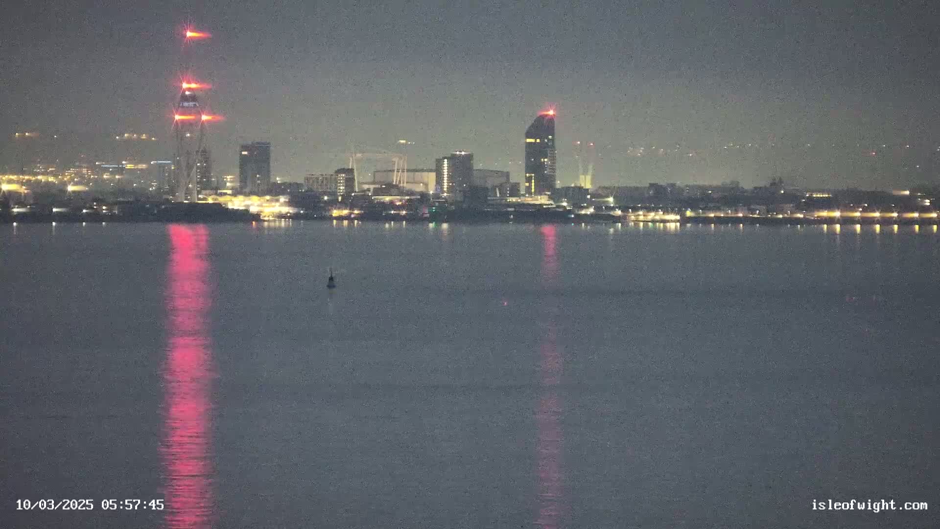 A nighttime view across calm water shows a city skyline with several illuminated buildings, including a tall tower with red lights, under a dark, clear sky.