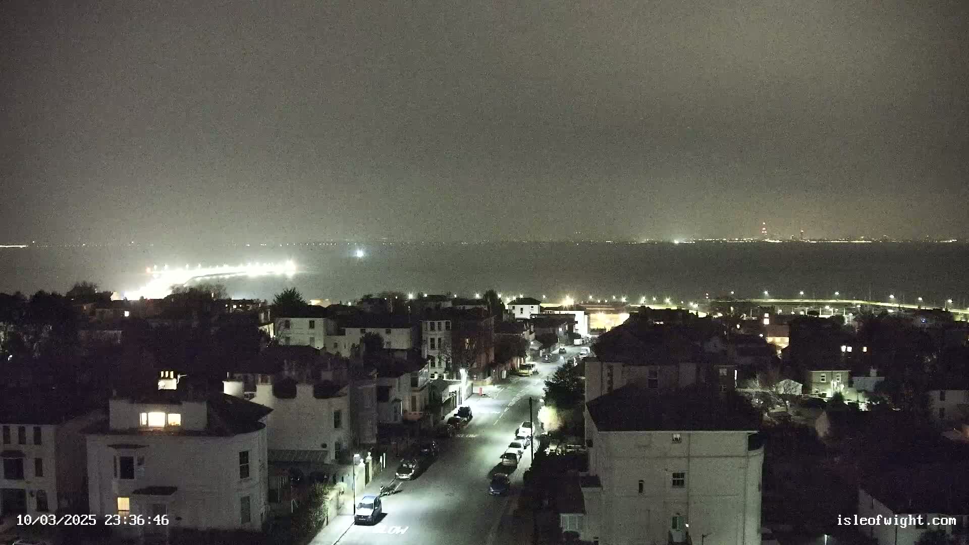 A nighttime view of a residential street overlooking a calm body of water with faintly visible lights across the horizon under a hazy sky.