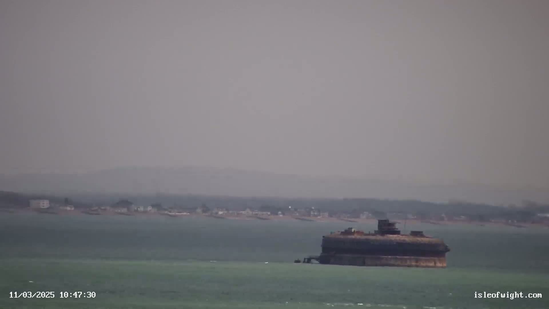 A hazy, overcast day reveals a large, dark structure resembling a fort in the calm, green-tinged water, with a distant shoreline visible in the background.